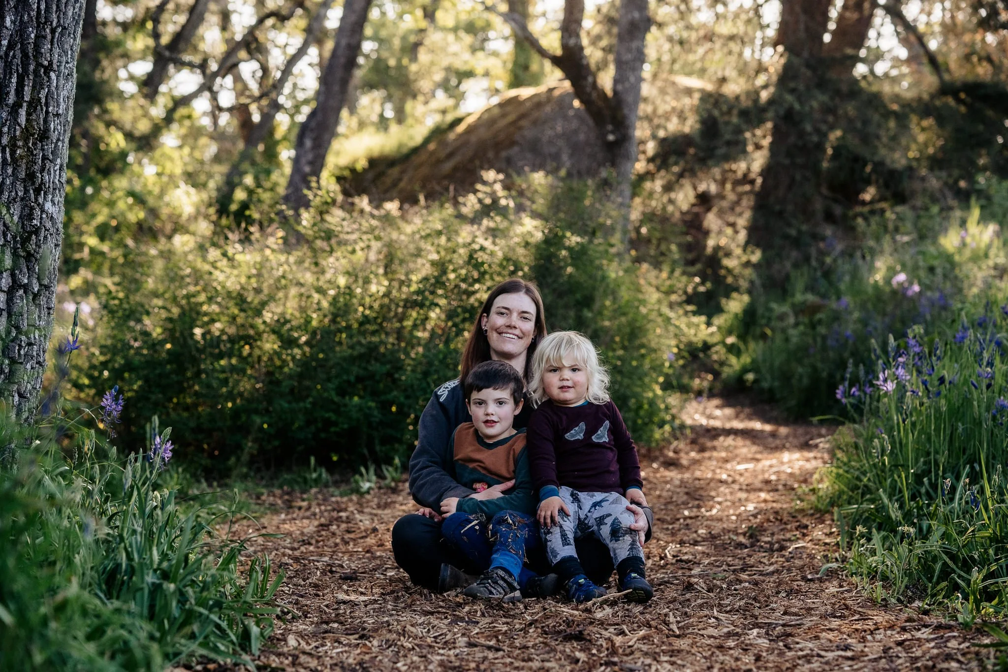 A woman sitting on a woodland path with two young children in front of her, surrounded by trees and greenery.