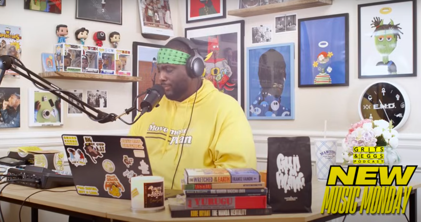 A man with a green headband and yellow hoodie speaking into a microphone with a laptop in front of him. The background is decorated with framed artwork, Funko Pop figures, and photos. Shirts and books are on the table, along with a floral arrangement and a clock.