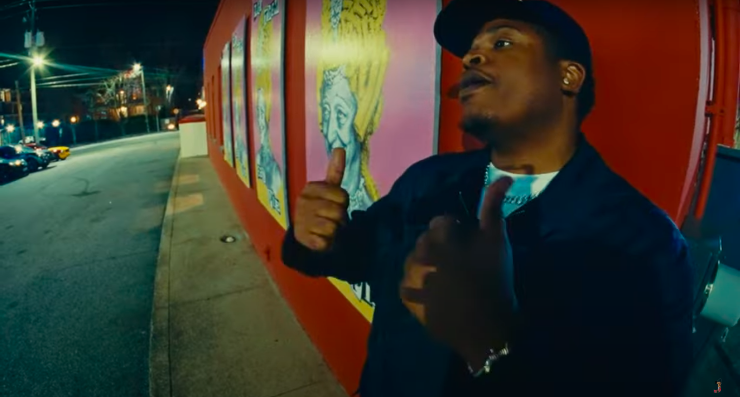 Man at night giving two thumbs up in front of colorful murals on a red wall.