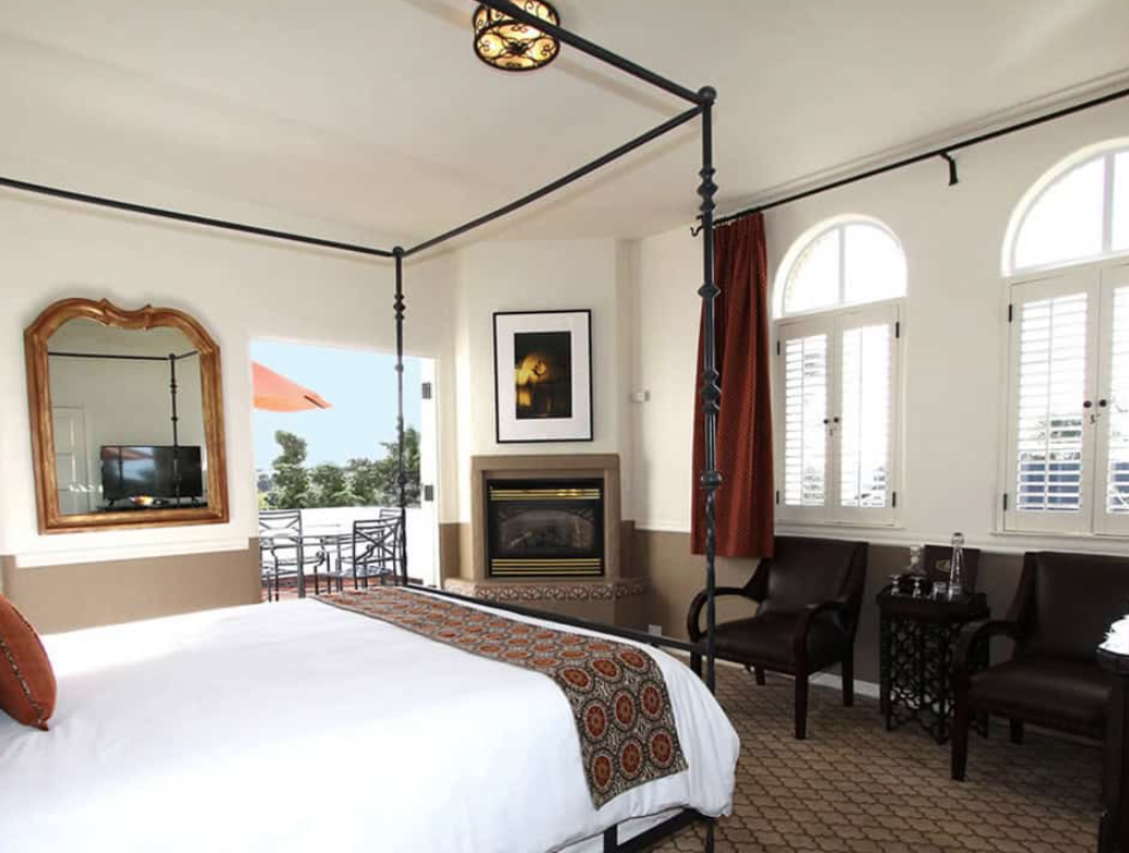 Hotel room with a four-poster bed, a fireplace, a flat-screen TV, large arched windows, and a balcony with outdoor seating.
