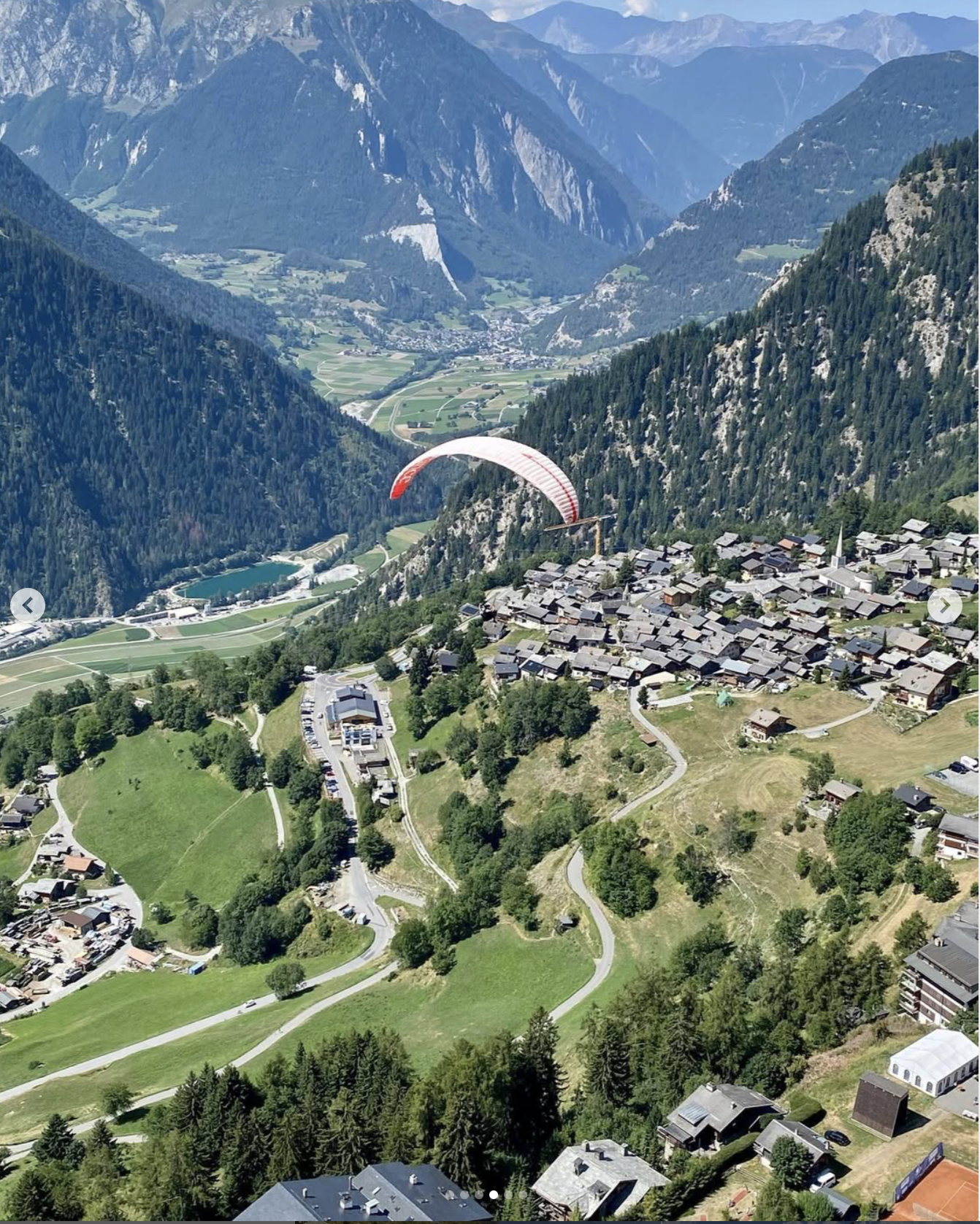 Aerial view of a small village nestled in the mountains with a paraglider flying above, surrounded by green hills, forests, and towering mountain peaks in the background in Verbier Switzerland near the W Verbier Hotel