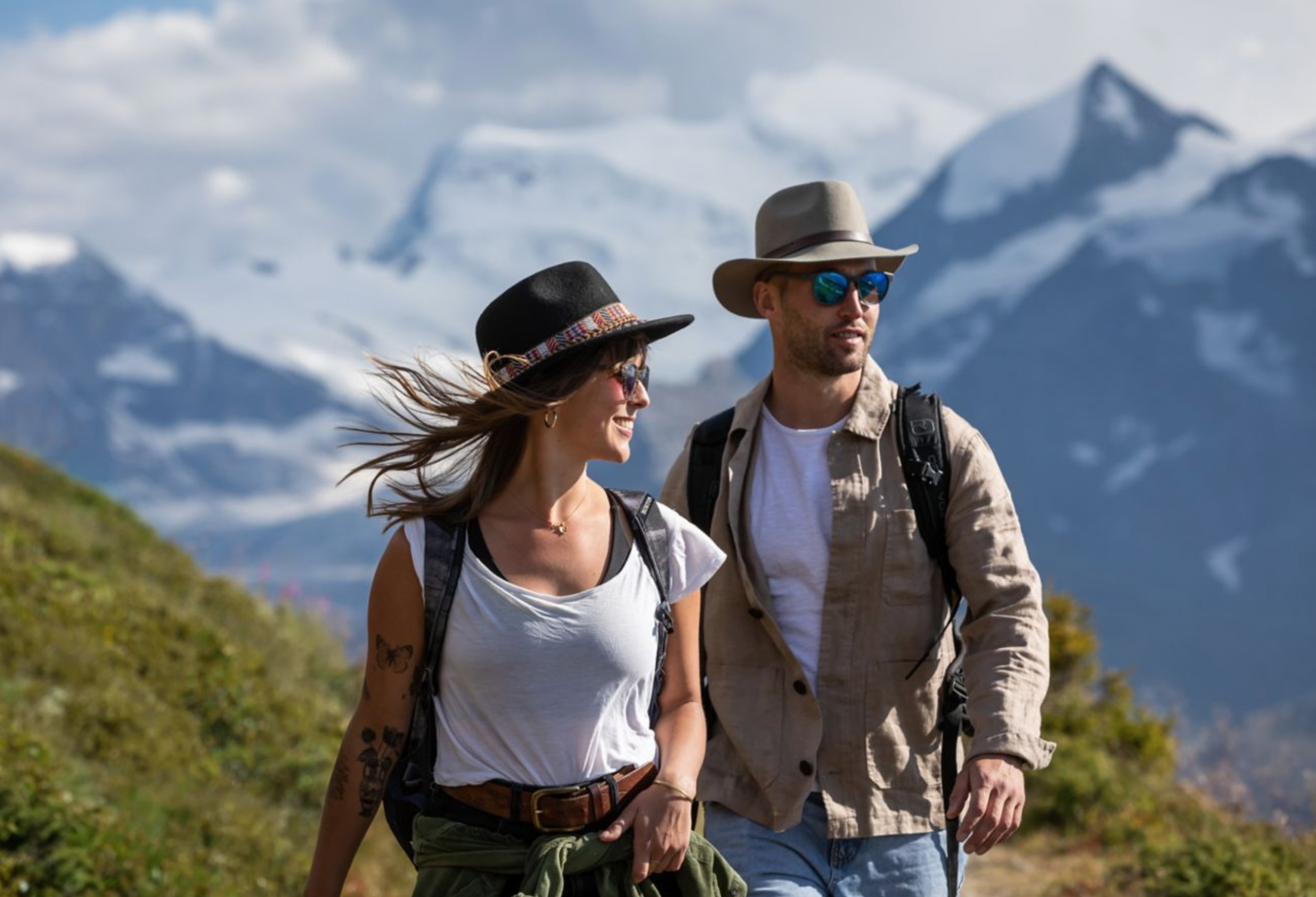 A smiling woman and man hiking outdoors in a mountainous area with snow-capped peaks in the background. They are wearing hats, sunglasses, and backpacks. Swiss Alps