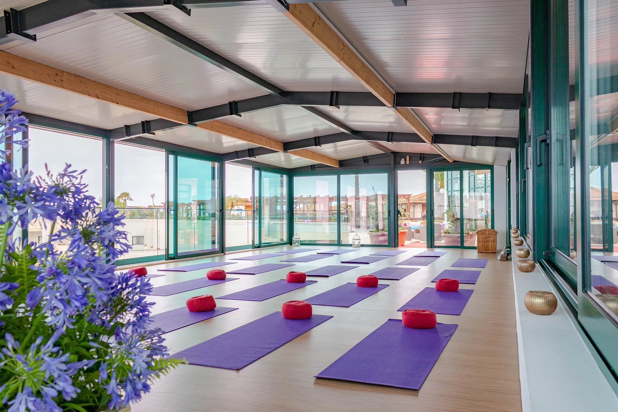 Interior of a bright yoga studio with purple mats and red meditation cushions, large windows, and wooden beams on the ceiling, with some plants and decorative stones.