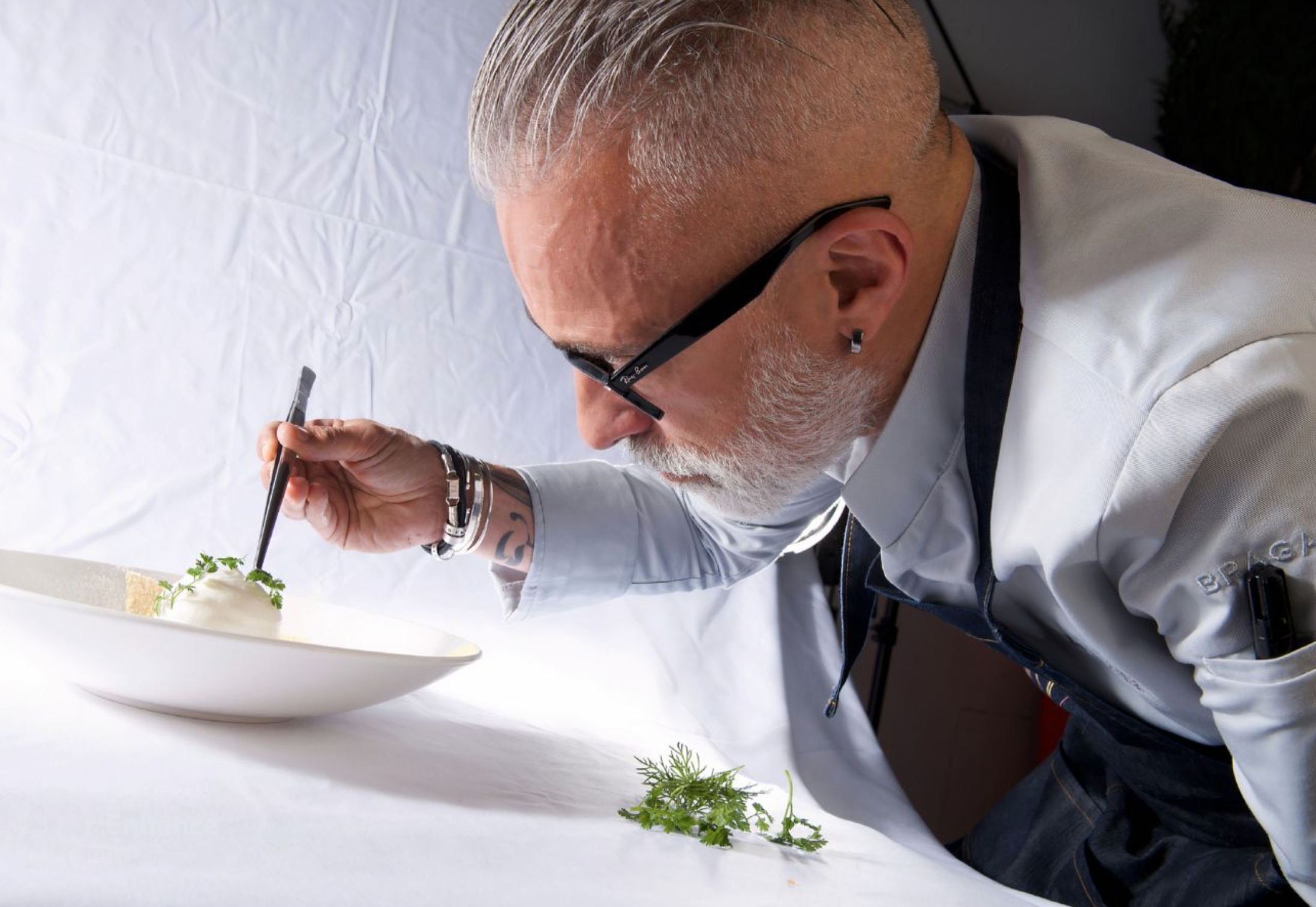 A man with gray hair and beard, wearing glasses and a chef's coat, is carefully decorating a dish with herbs using tweezers in a photography studio. W Verbier, Switzerland Michelin