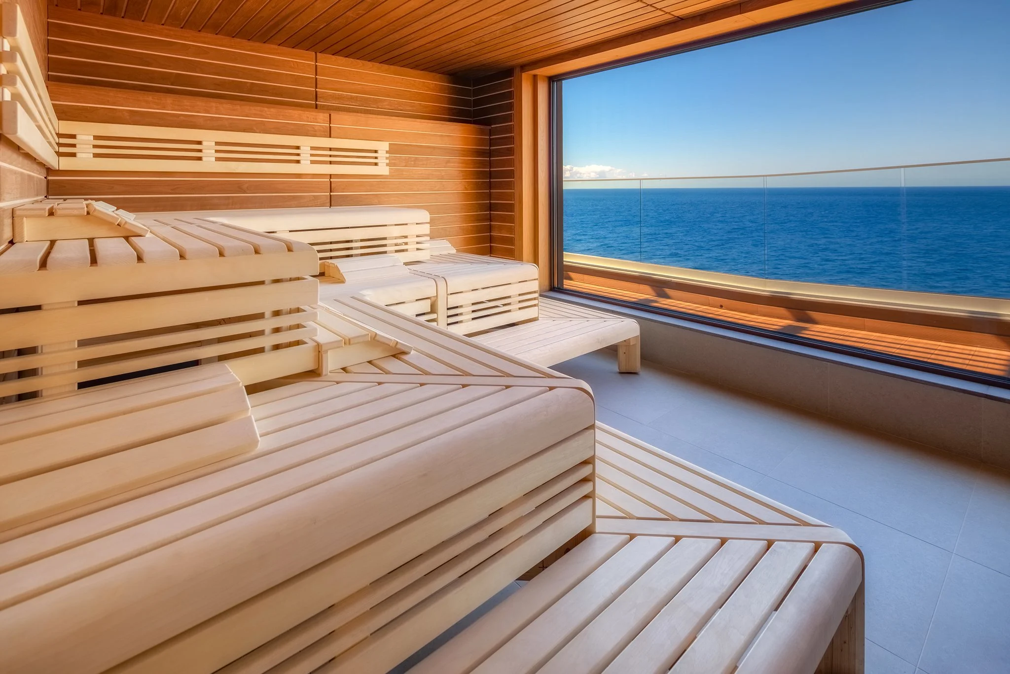 A wooden sauna room with benches and a large glass window offering a view of the ocean and a blue sky.