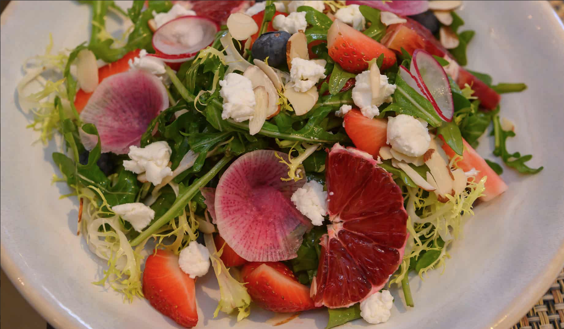 Close-up of a fresh mixed fruit and salad with sliced strawberries, grapefruit, watermelon, blueberries, radish slices, arugula, frisée lettuce, crumbled feta cheese, and sliced almonds on a white plate.
