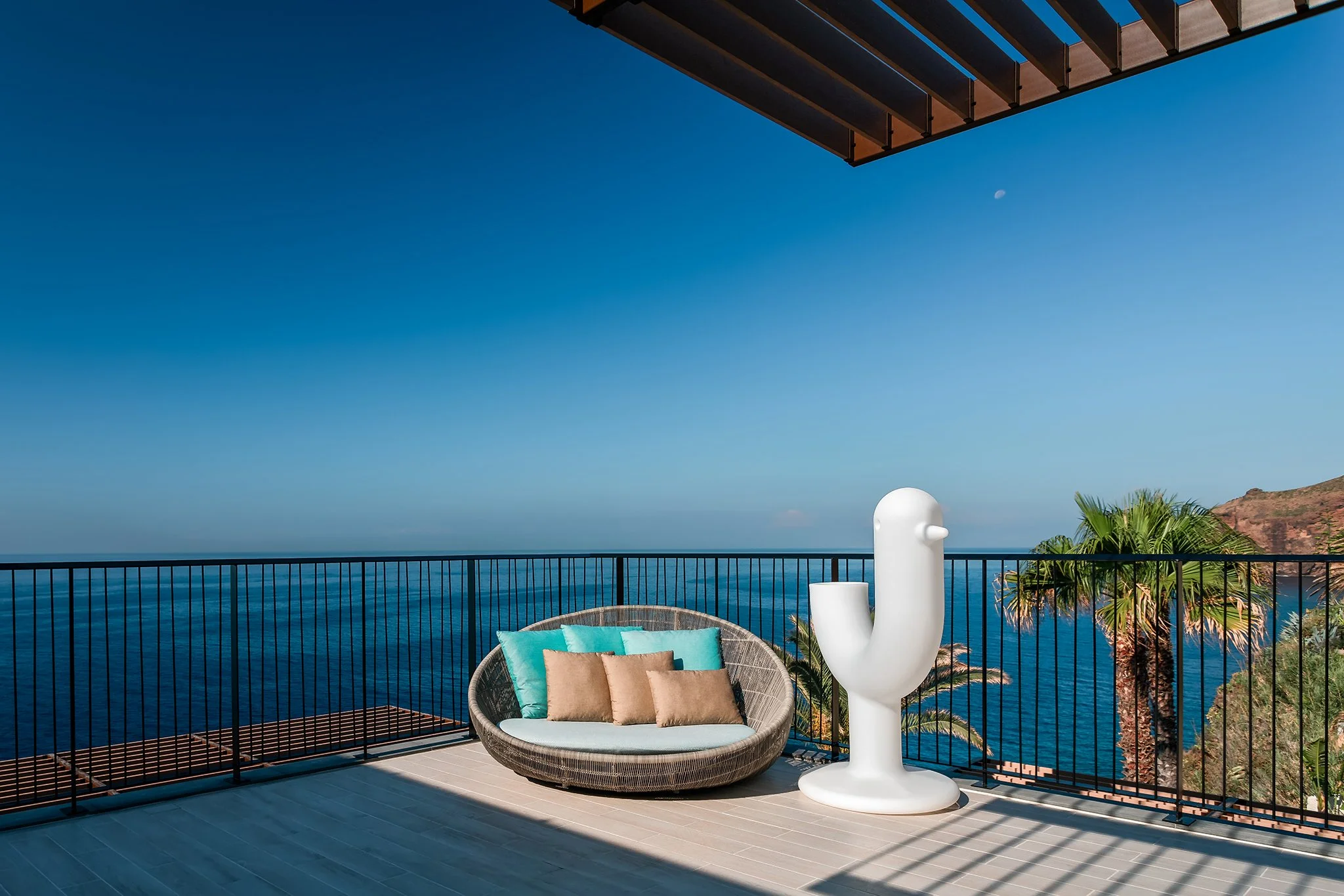 Balcony overlooking the ocean with a round wicker chair with turquoise and beige pillows, a white sculpture resembling a bird, a palm tree, and a view of cliffs in the distance under a clear blue sky.
