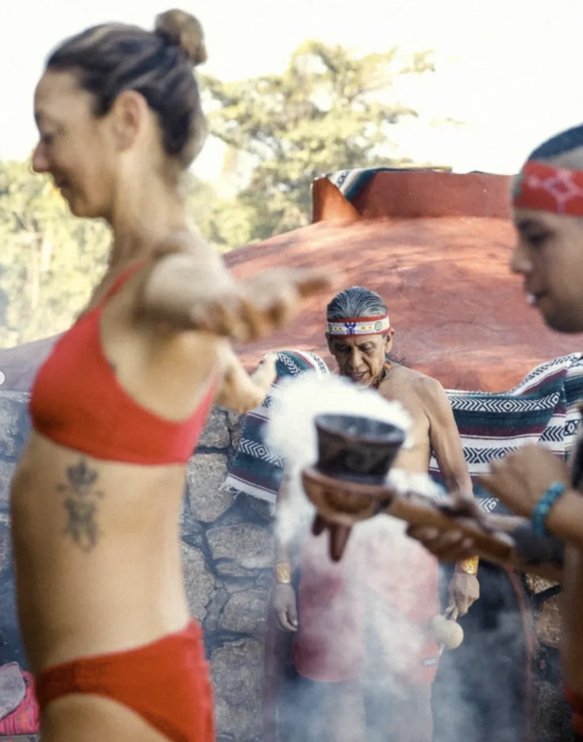 Indigenous man sitting outdoors, surrounded by women and children dressed in traditional attire, with a large clay pot behind him, in a rural setting with trees. Temescal Xinalani