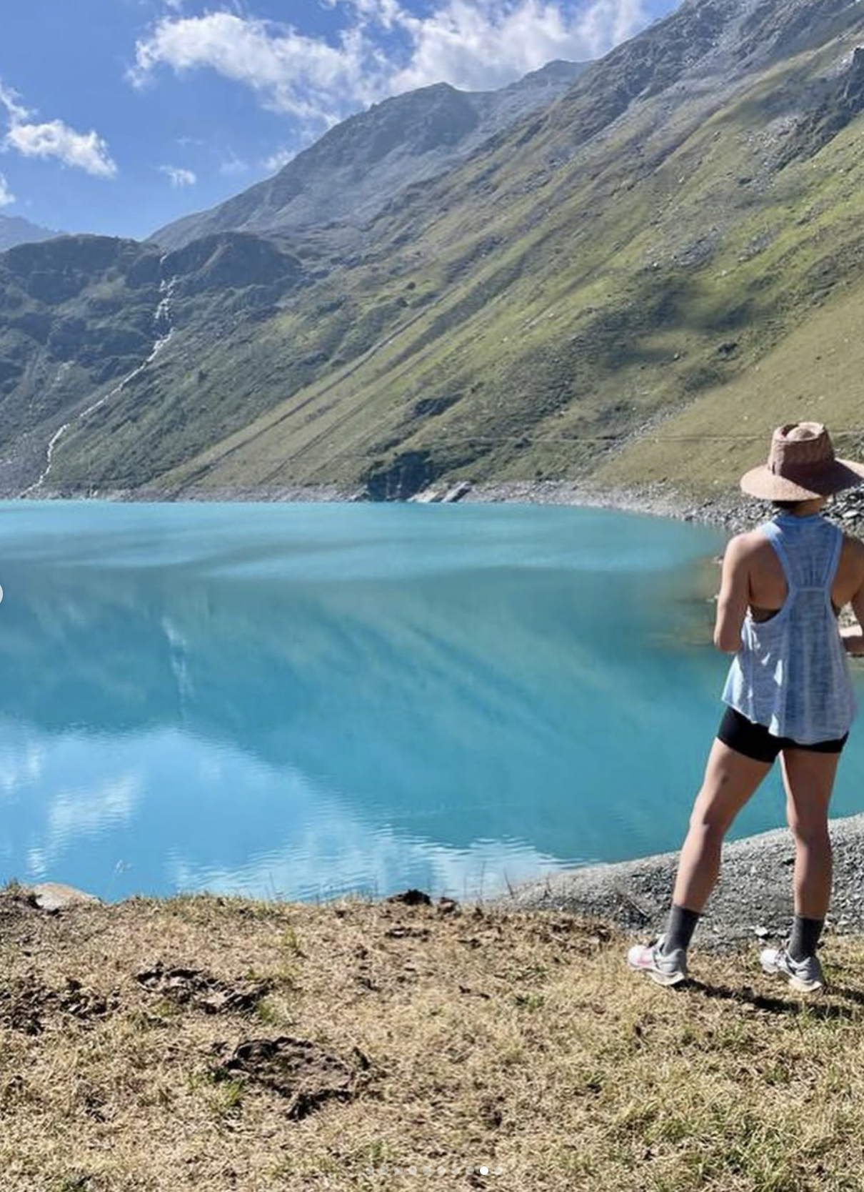 A person wearing a wide-brimmed hat, sleeveless top, shorts, and sneakers standing by a bright blue lake, with mountains in the background under a partly cloudy sky.