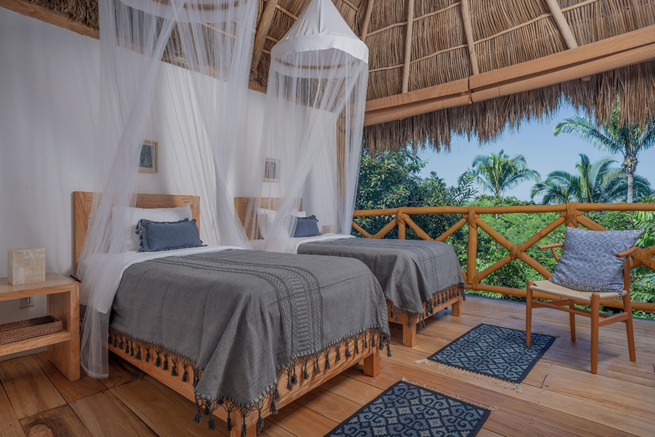 Description A tropical bedroom with a thatched roof, a large bed with striped pillows, hanging wicker light fixtures, a small wooden side table with a cup, and a balcony with a hammock and view of green trees and blue sky. Bookyogaretreatsgoes here