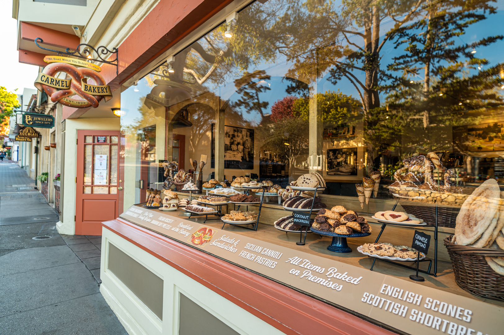 Bakery display window with assorted baked goods including cookies, croissants, muffins, and brownies. Exterior of Carmel Bakery with signage and sidewalk outside on a sunny day.