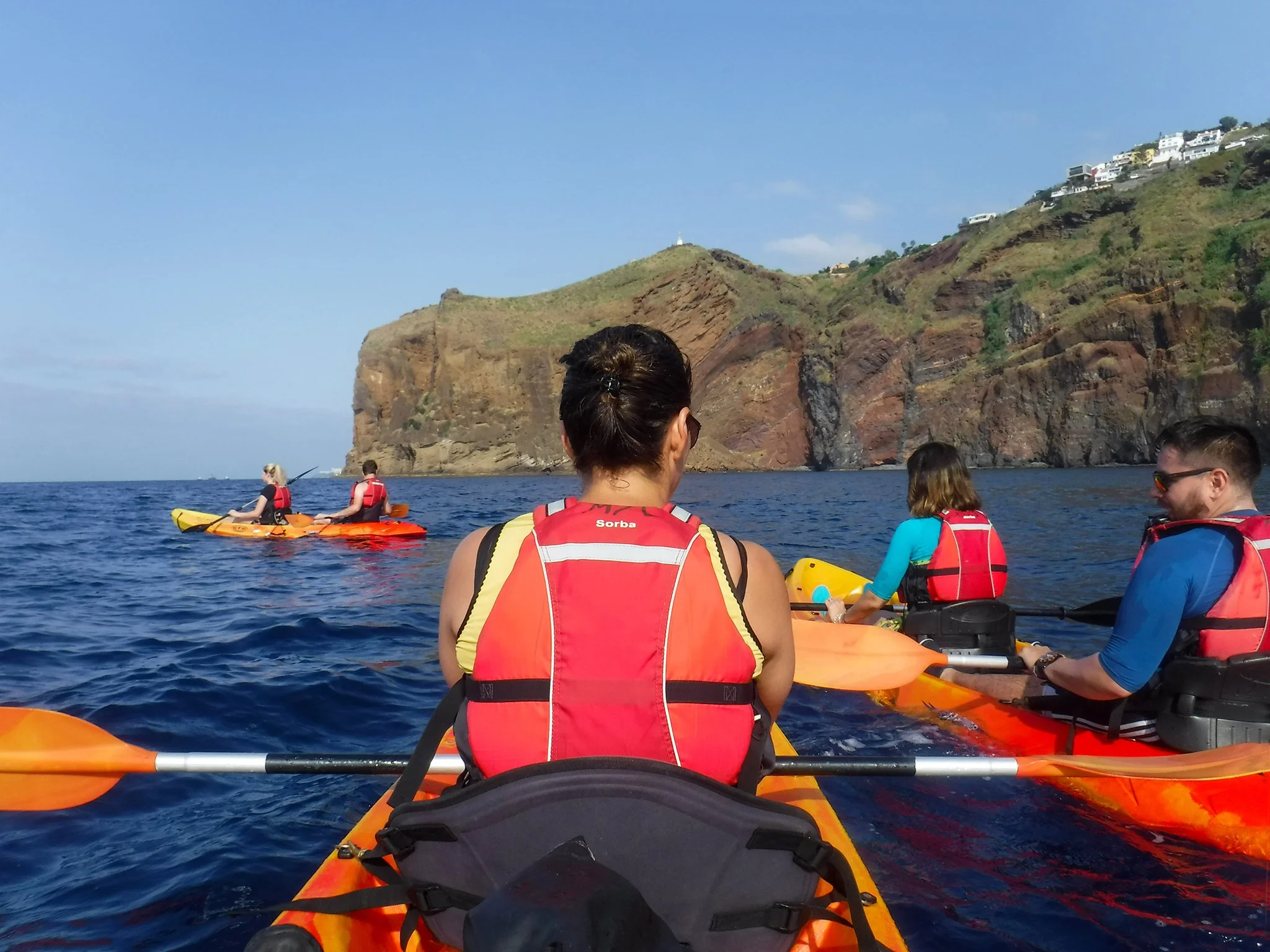 People kayaking in the ocean near rocky cliffs and hillside homes.