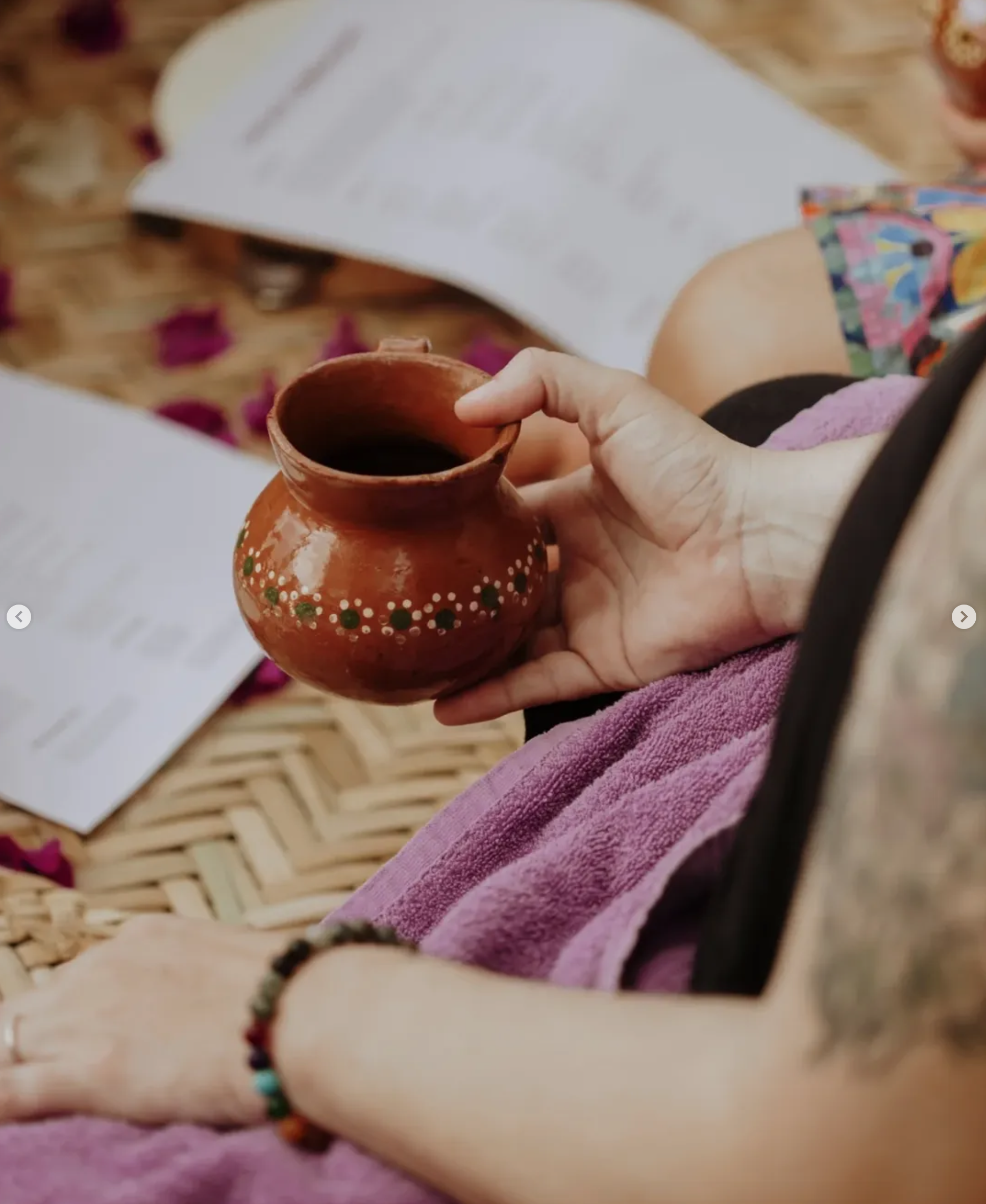 A person holding a small, traditional clay cup with a painted dotted design, filled with a dark beverage, sitting on a woven mat with scattered flower petals and papers around. Cacao Ceremony_Xinalani