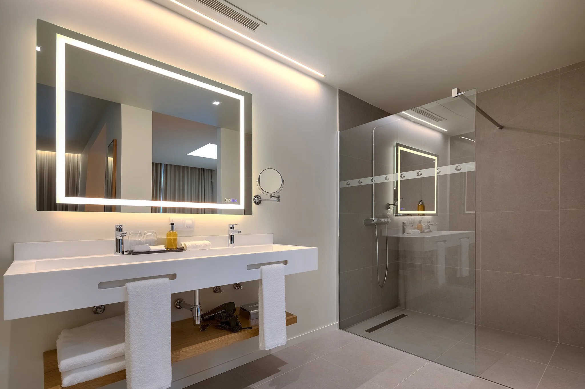 Modern bathroom with large illuminated mirror above a double sink vanity, glass shower enclosure, and minimalist decor.