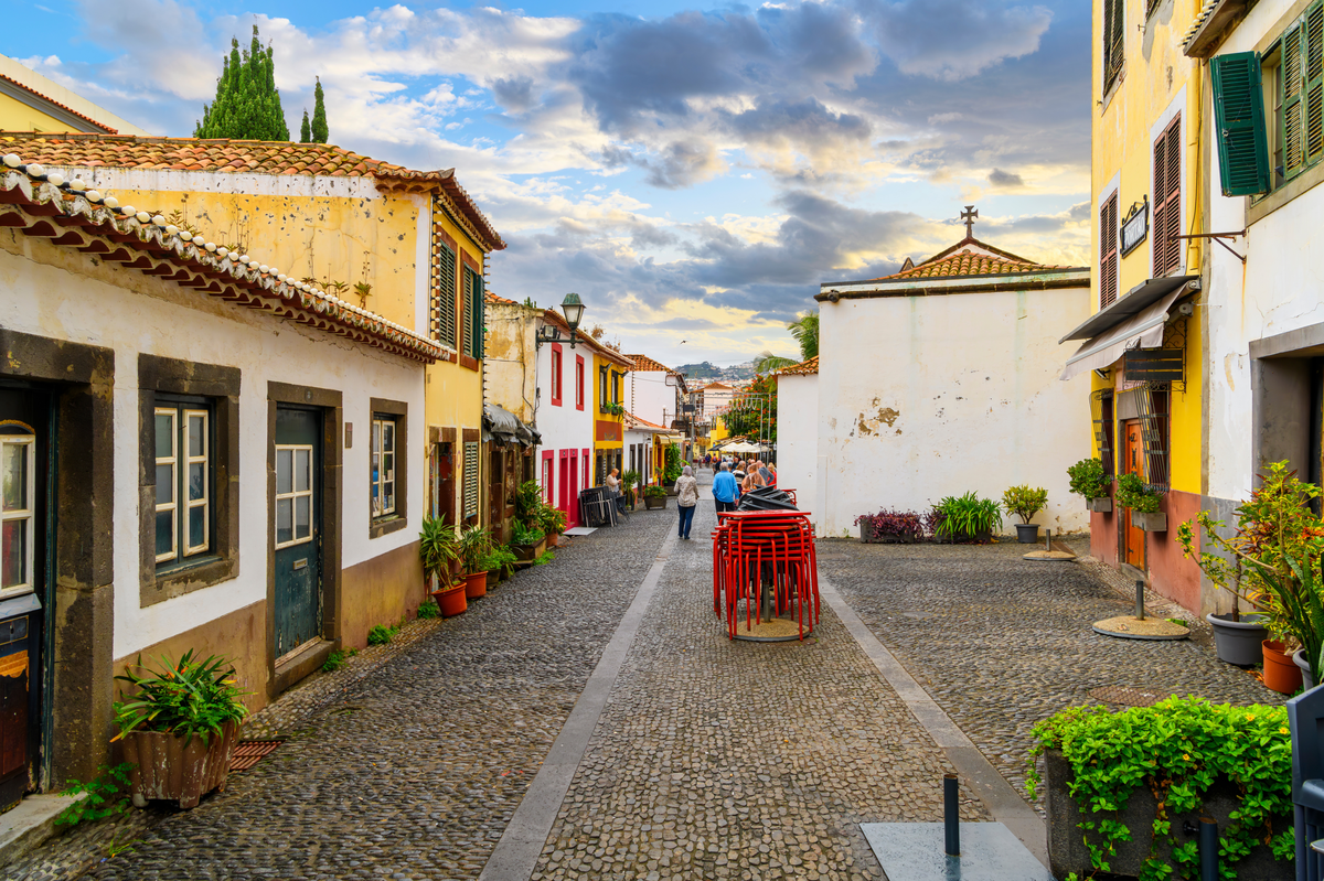 A narrow cobblestone street in a European town, lined with colorful, old buildings with green shutters and potted plants. People are walking and sitting at outdoor cafes beneath a partly cloudy sky.