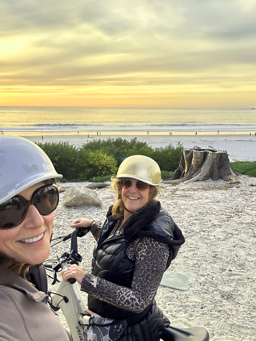 Two women wearing helmets and sunglasses smiling while riding bikes on a beach at sunset, with a large tree stump and lush greenery around them and the ocean in the background.