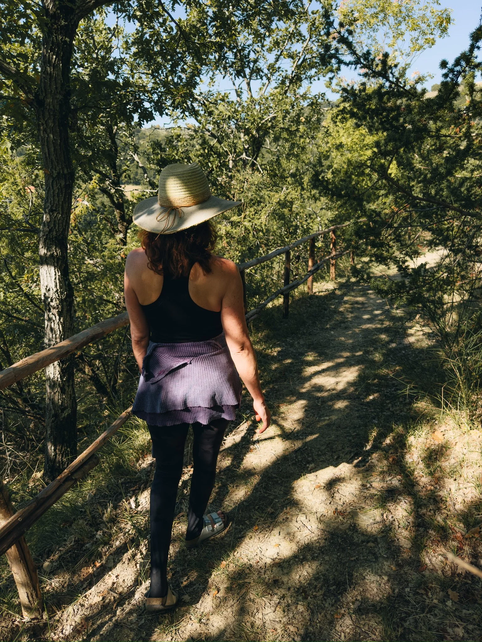 Woman walking on a dirt trail through a wooded area, wearing a sun hat, black top, patterned skirt, black tights, and sandals.