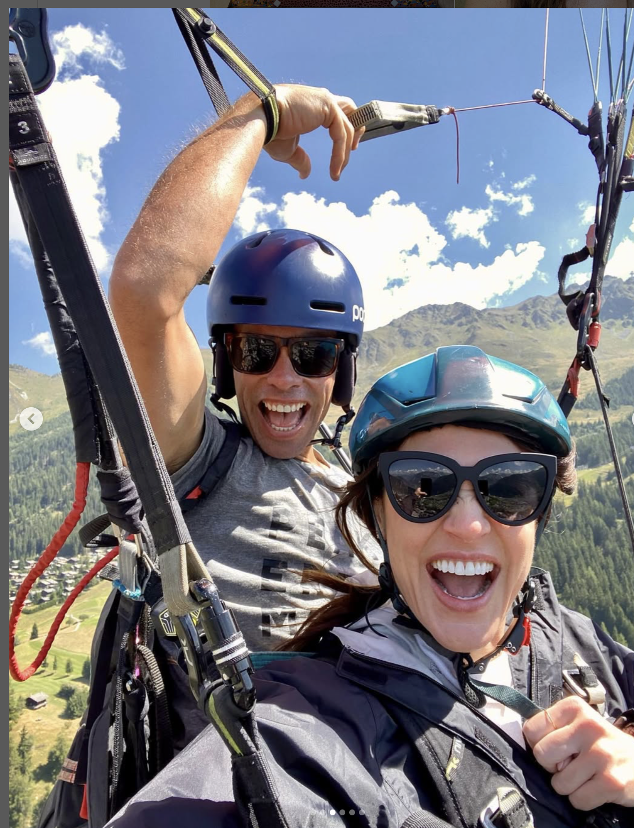 Two people wearing helmets and sunglasses paragliding over a mountainous landscape with green hills and scattered houses under a blue sky.