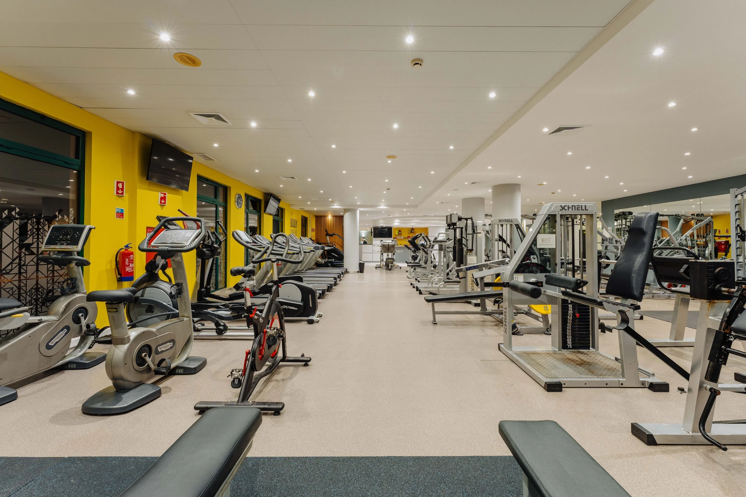 A spacious indoor gym with various exercise equipment including treadmills, stationary bikes, and weight machines, with yellow walls and a white ceiling with bright lighting.