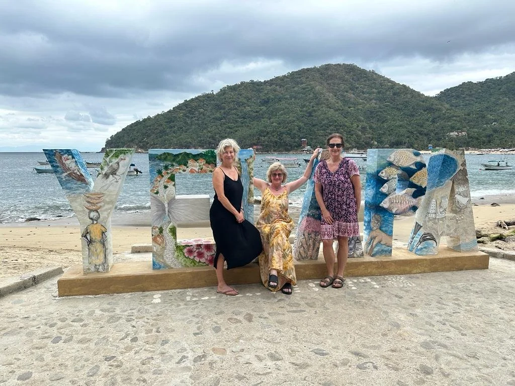 Three women standing and sitting in front of large painted letters spelling 'VEMA' on a beach, with boats in the water and a hilly landscape in the background on a cloudy day.