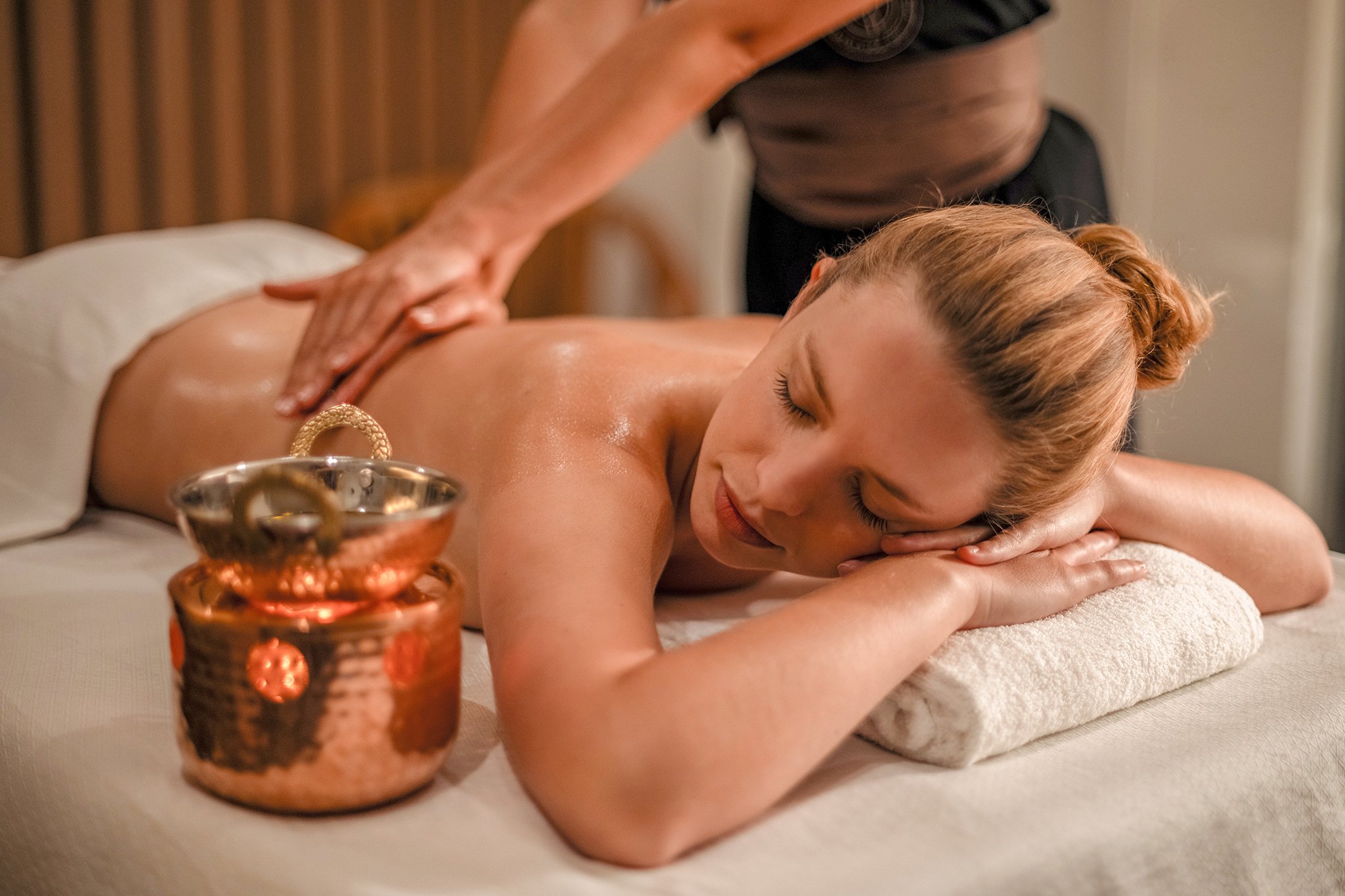 A woman with red hair receives a massage from a massage therapist in a spa setting, with a copper-colored candle holder in the foreground.