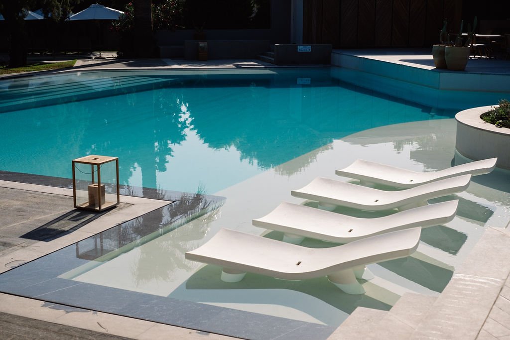 Empty swimming pool with four white lounge chairs partially submerged, a small side table, and potted plants in an outdoor setting.