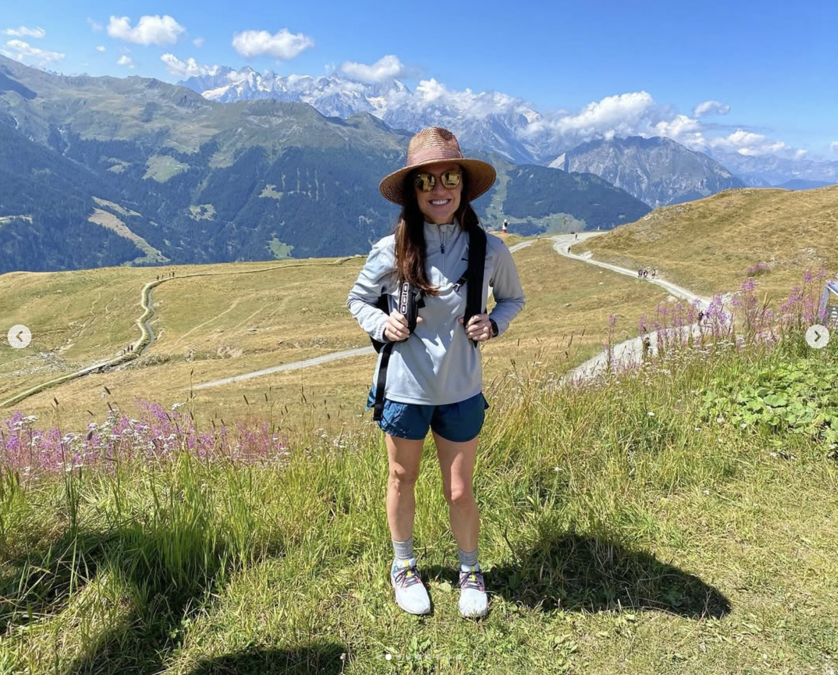 A woman hiking in a mountainous area, wearing a wide-brimmed hat, sunglasses, a light-colored jacket, shorts, and hiking shoes, with a backpack, standing on a grassy trail with mountains and a partly cloudy sky in the background.