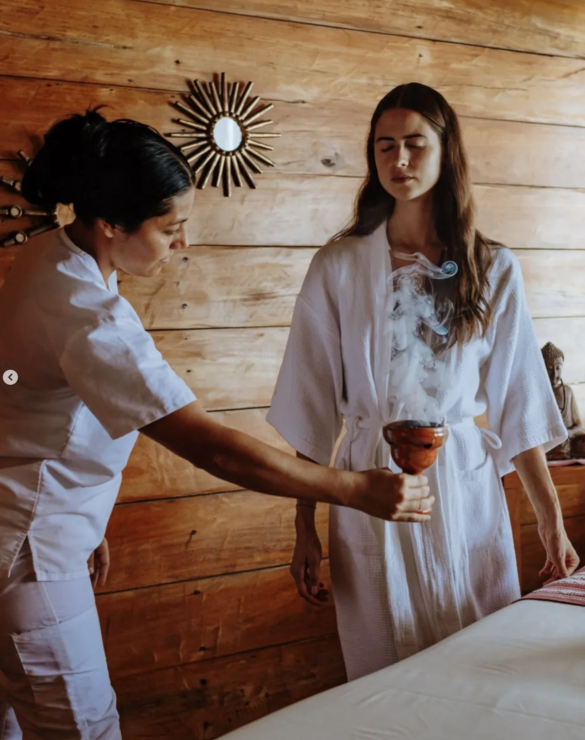 A woman in a white bathrobe receiving a smudging ritual from another woman in a white outfit in a room with wooden walls. Massage_Xinalani