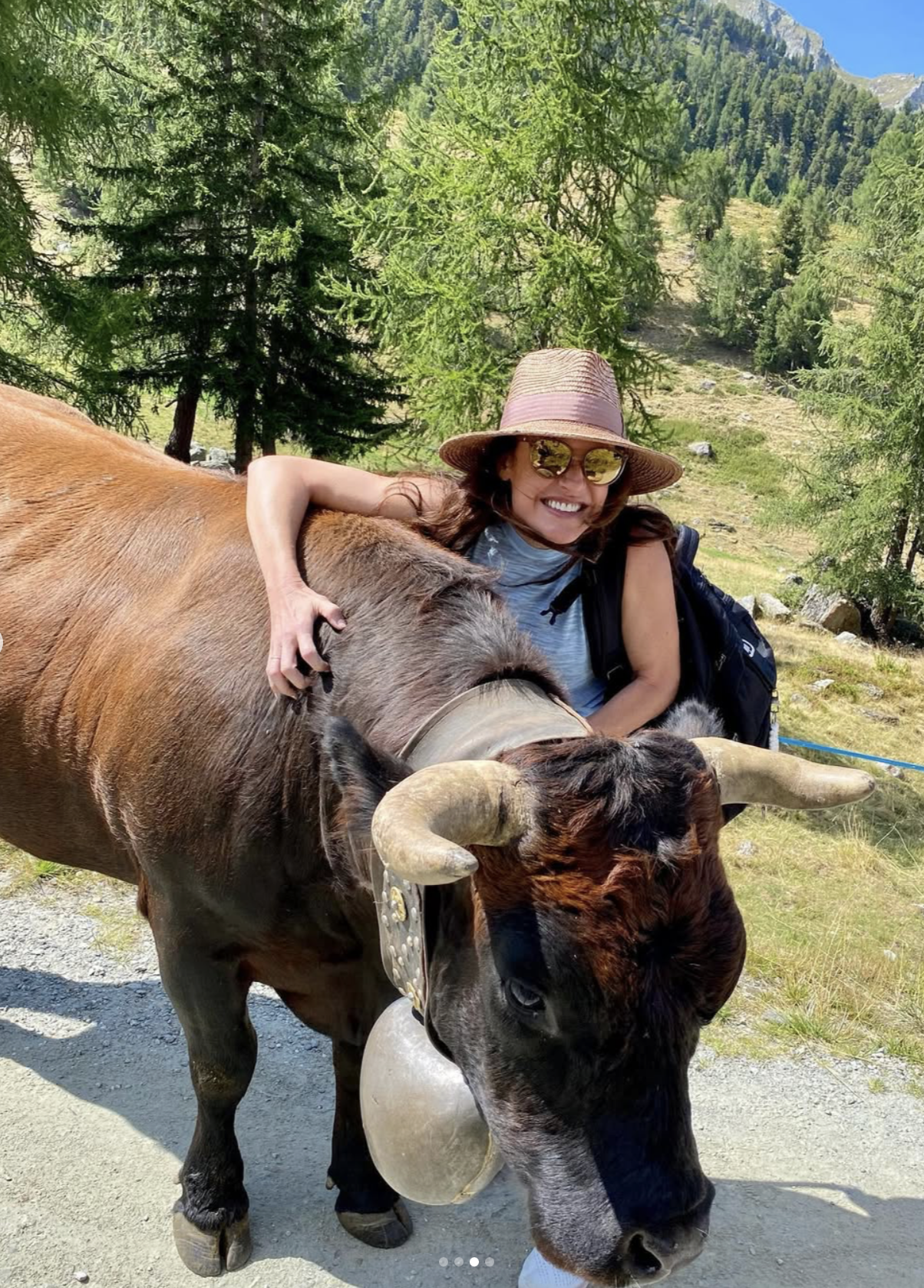 Melissa Barron, woman with a sunhat and sunglasses hugging a large brown cow with big horns, in a green forested mountain area on a sunny day in Verbier Switzerland