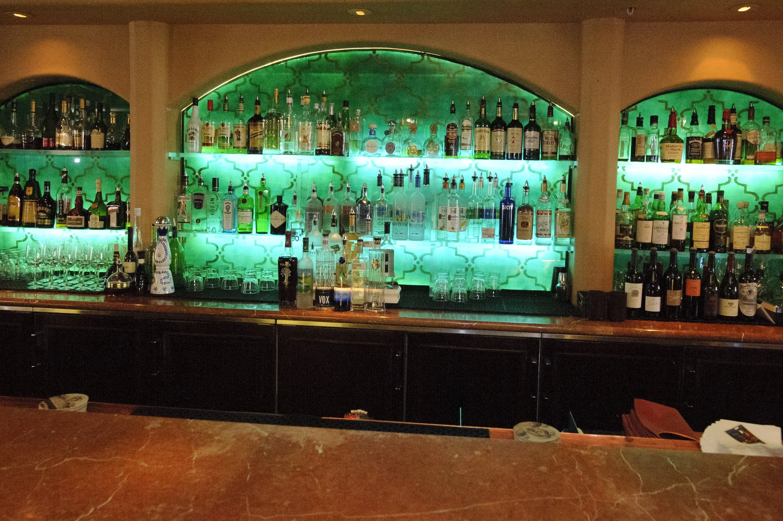 Backlit bar display with various bottles of alcohol, shot glasses, and bar tools on a green patterned background.