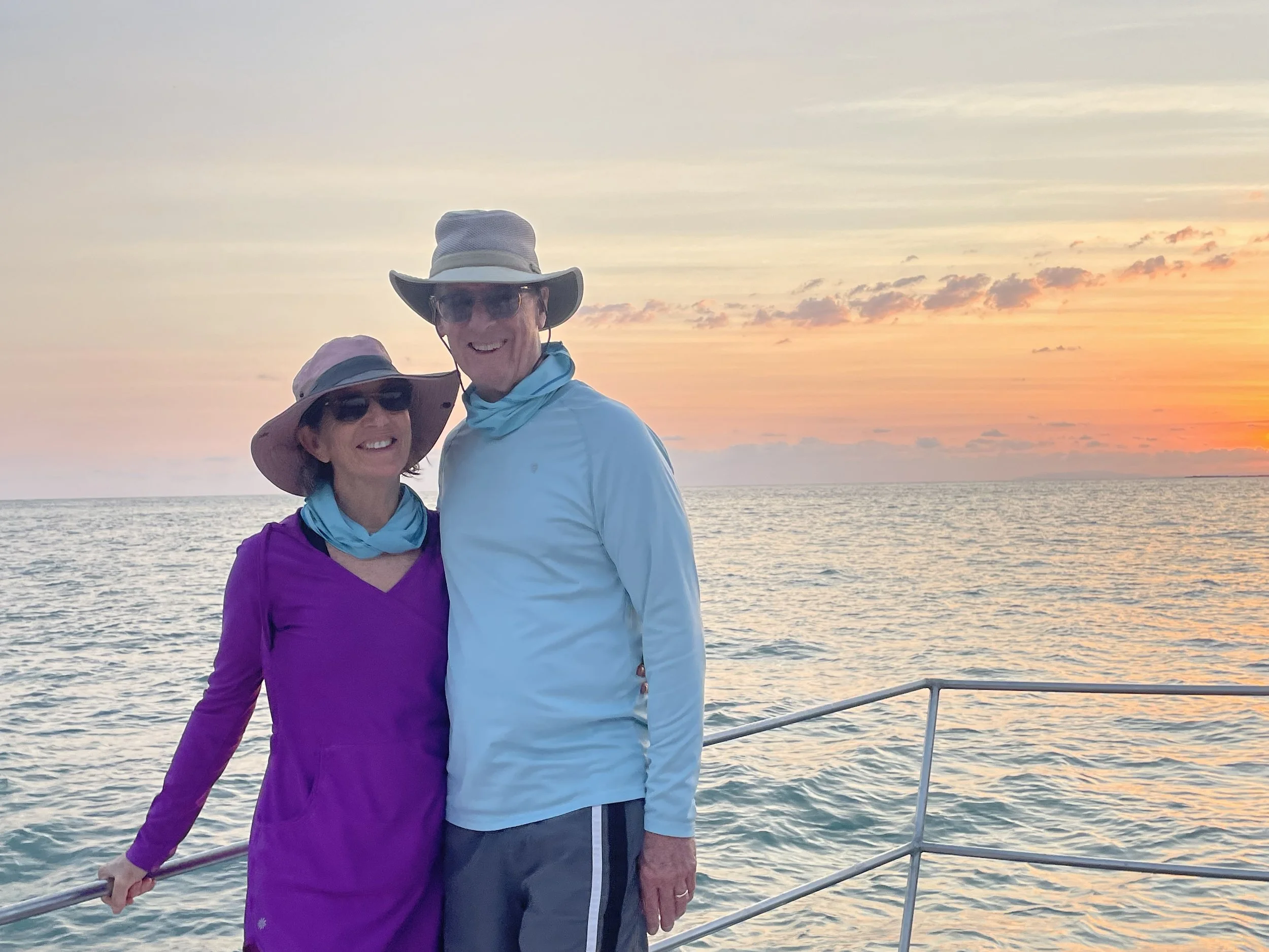 Two people, a man and a woman, smiling on a boat during sunset with ocean in the background. They are wearing hats and sunglasses, dressed in casual outdoor clothing.