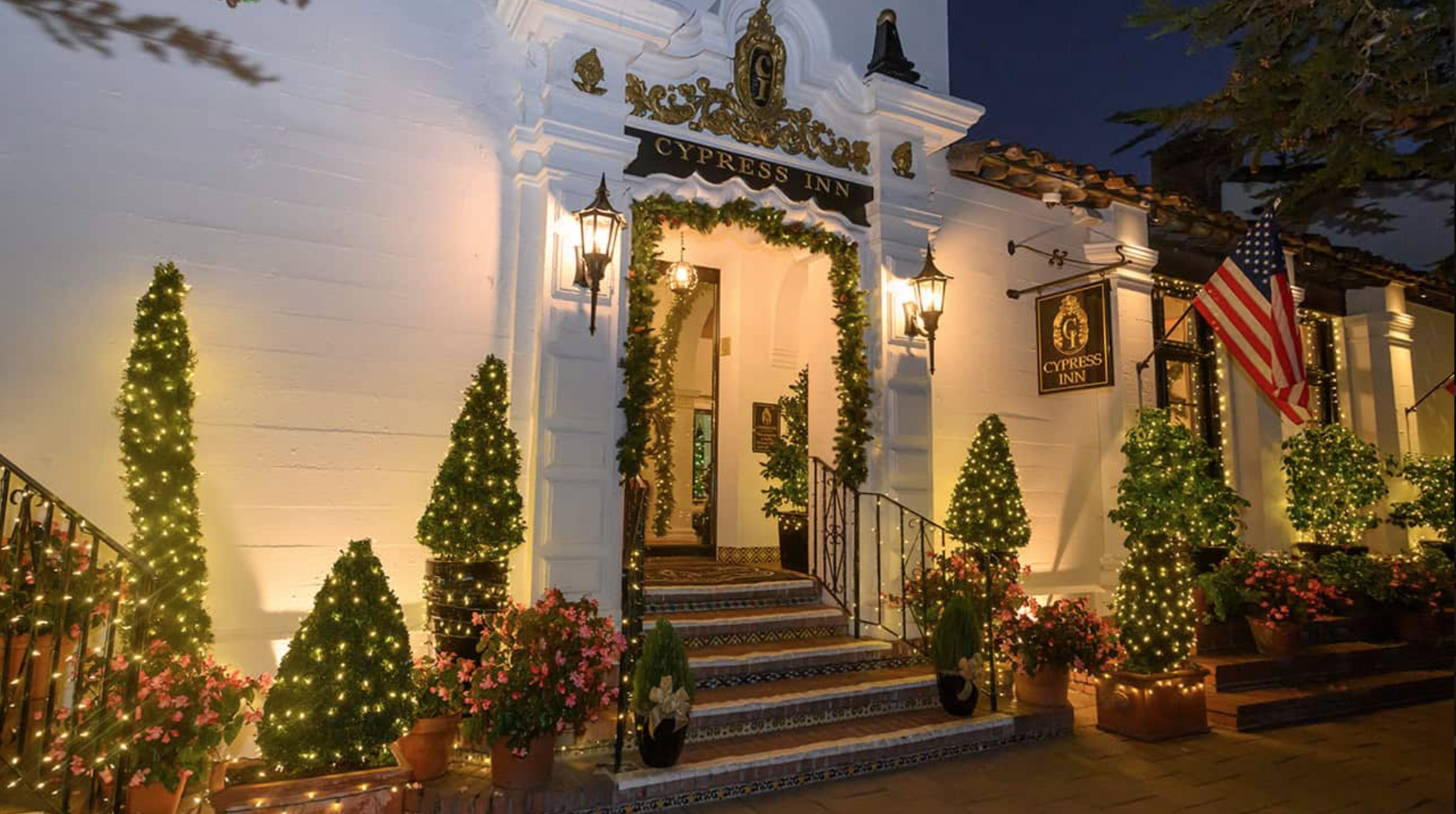 The exterior of Cypress Inn decorated with Christmas lights, garlands, and potted poinsettias, with an American flag, during the evening.