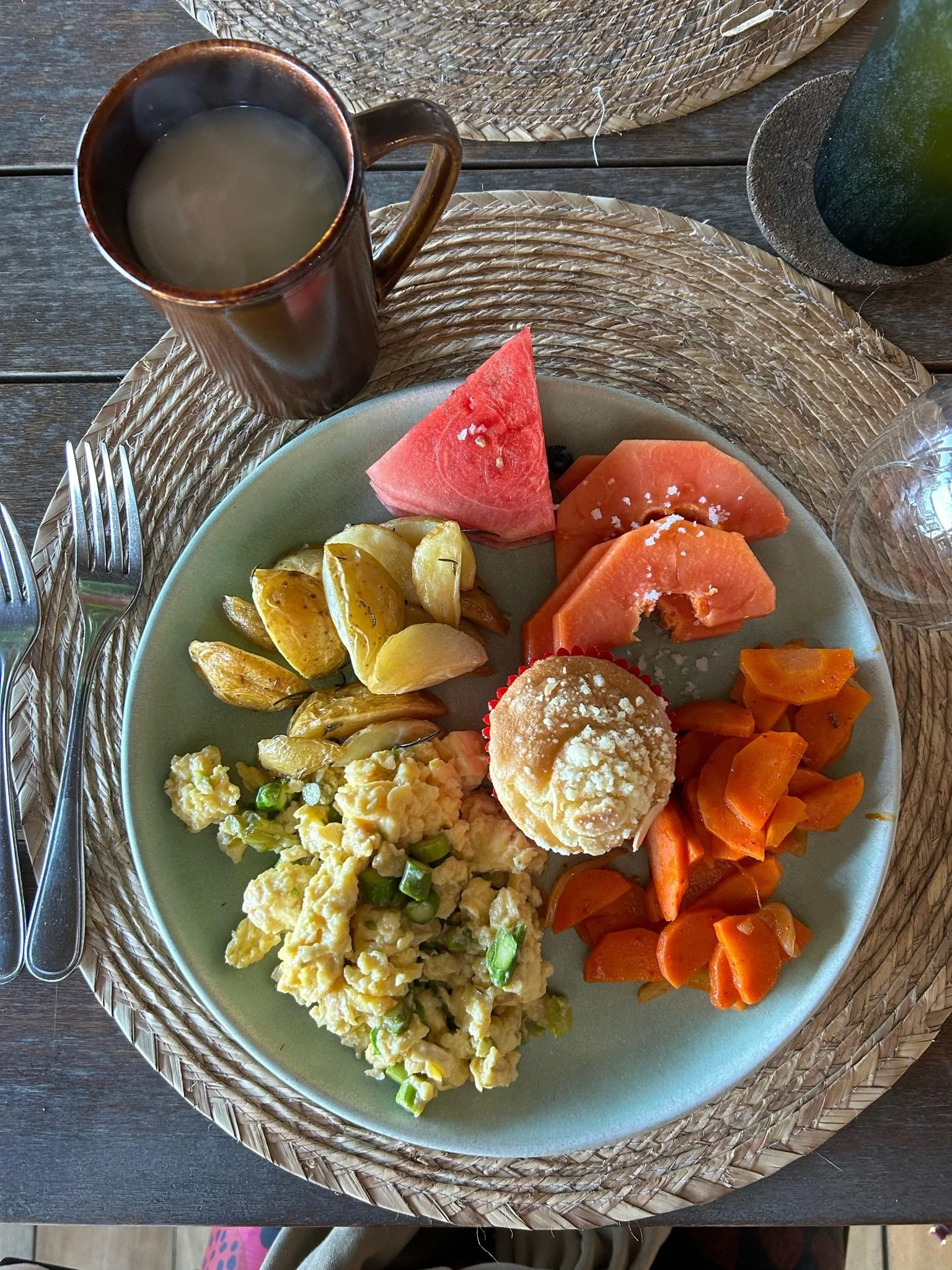 A breakfast plate with scrambled eggs, roasted potatoes, sliced carrots, watermelon, papaya, and a muffin, with a mug of coffee and an empty glass on a woven placemat.