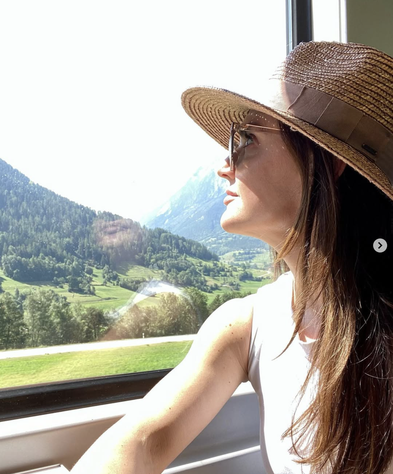 Woman wearing a large straw hat and sunglasses looking out the window at a scenic mountain landscape with green hills and trees.