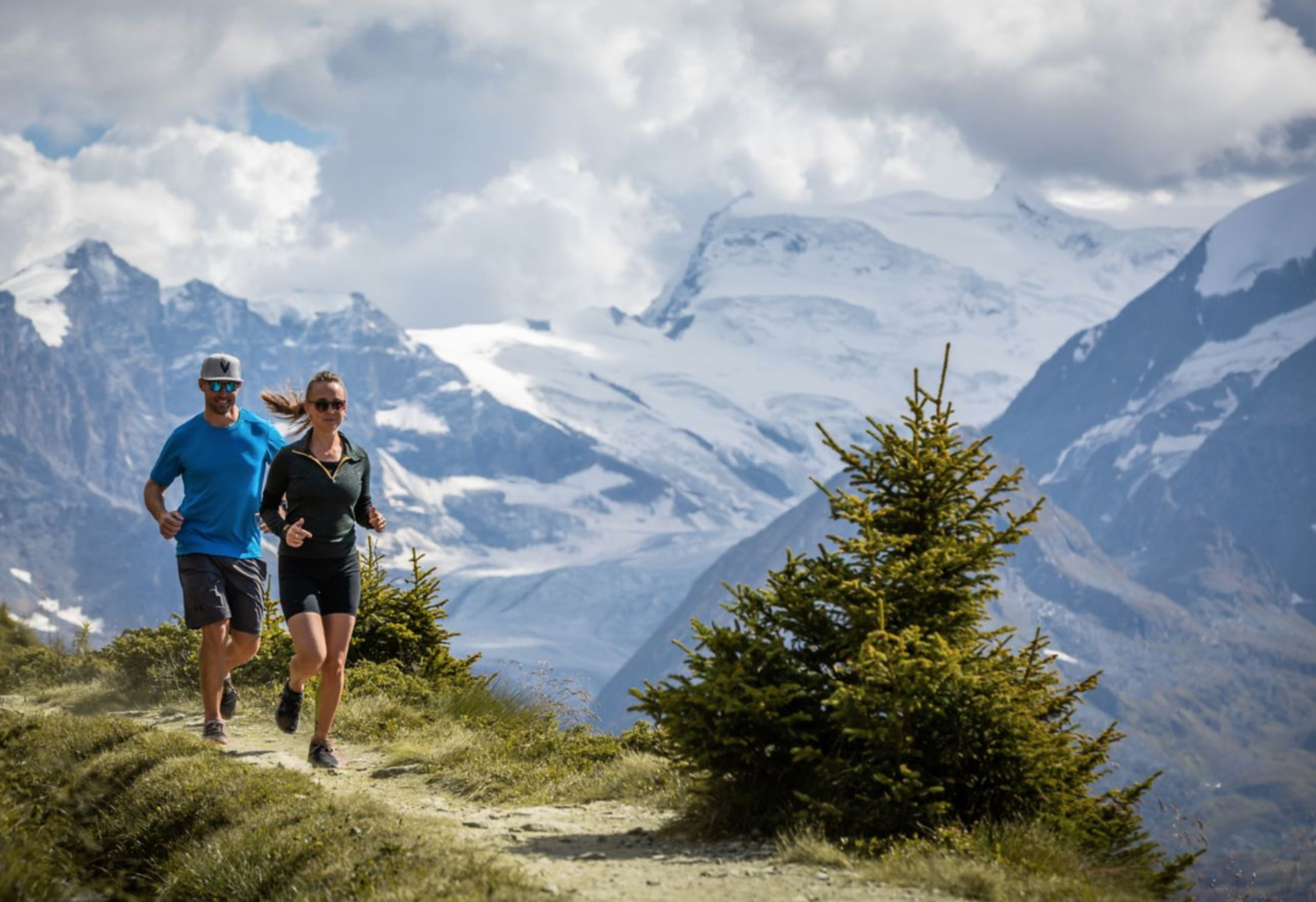 Two people running on a trail in a mountainous area with snow-capped peaks and trees under a partly cloudy sky. Swiss Alps