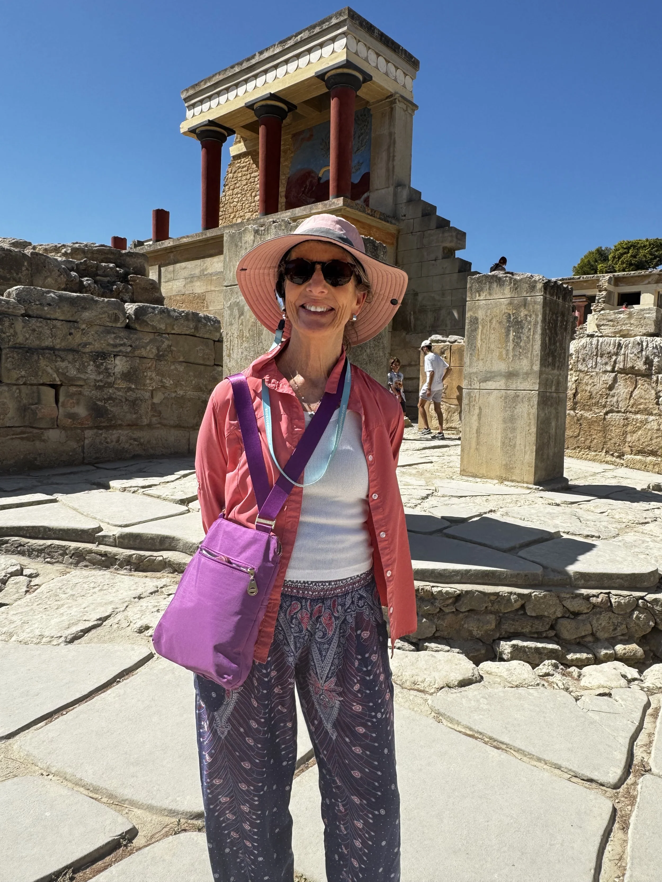 A smiling woman, Joanna Brown, yoga instructor, wearing patterned pants and a pink jacket, standing outdoors near ancient stone ruins with steps, a partially restored building, and a clear blue sky in the background.