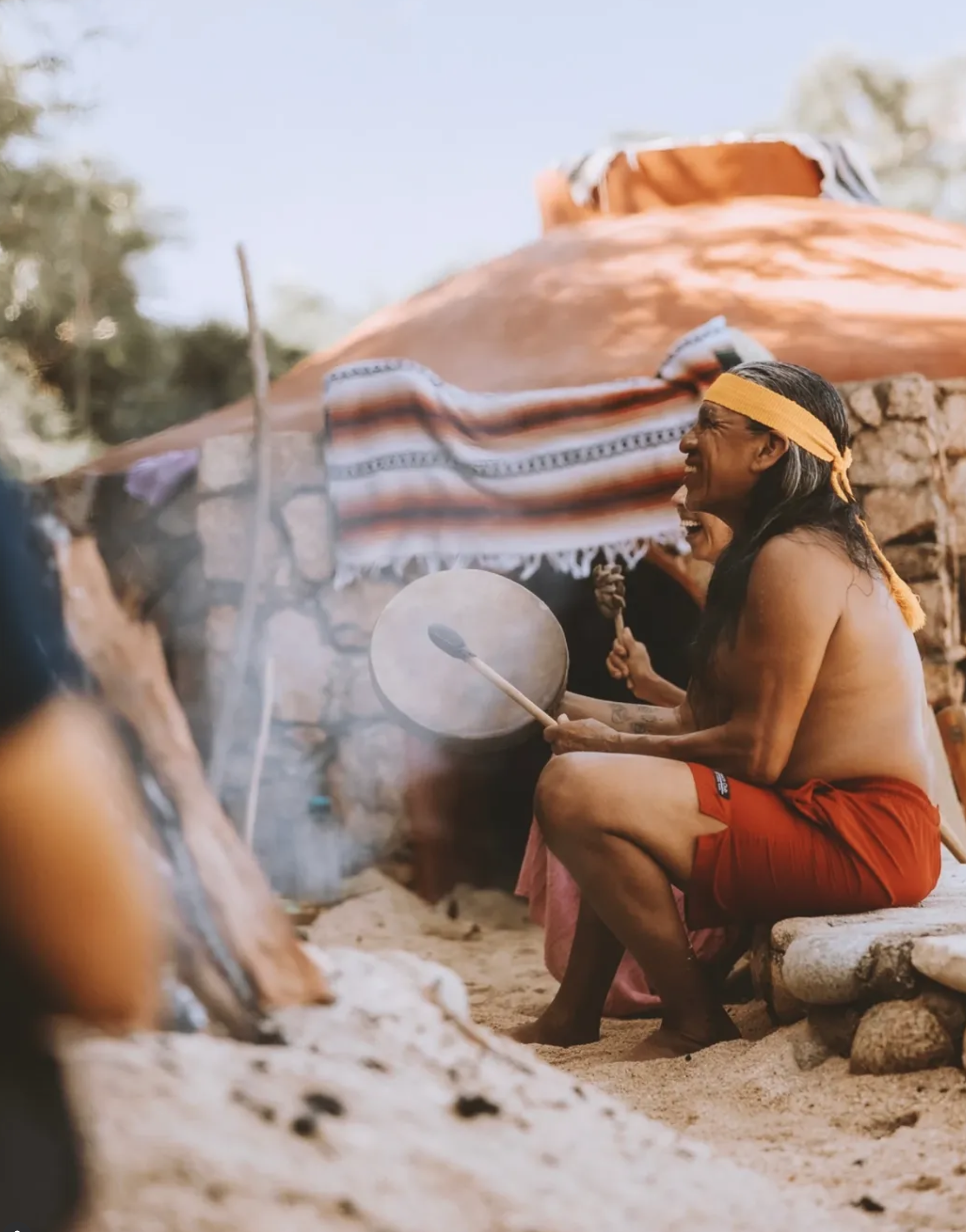 An Indigenous man sitting on a stone bench outside a traditional round hut, playing a drum and laughing, with a colorful cloth hanging on the hut and trees in the background.