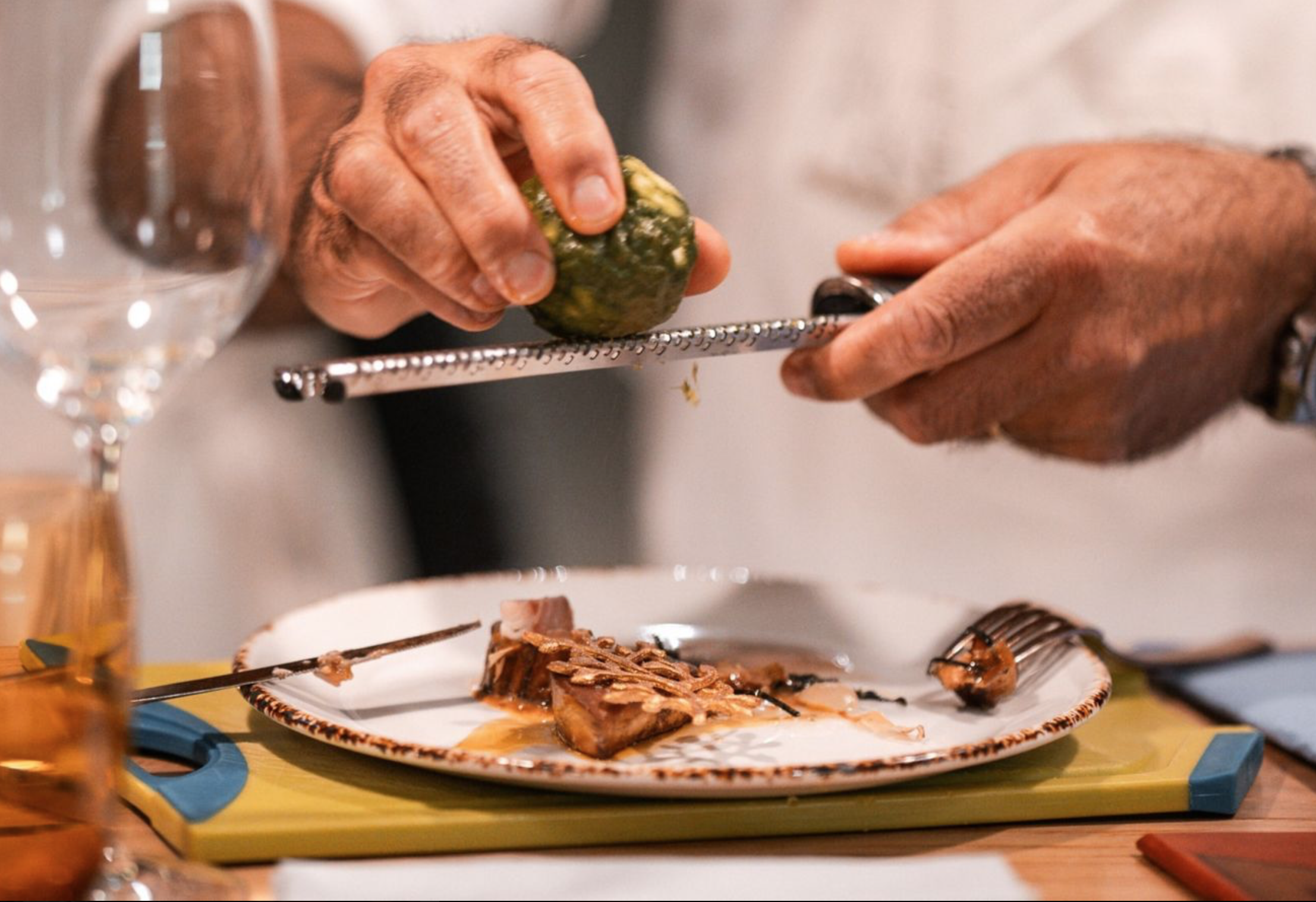 A person grating lemon or lime over a plate with cooked meat slices, surrounded by glasses and kitchen utensils. W Verbier Michelin