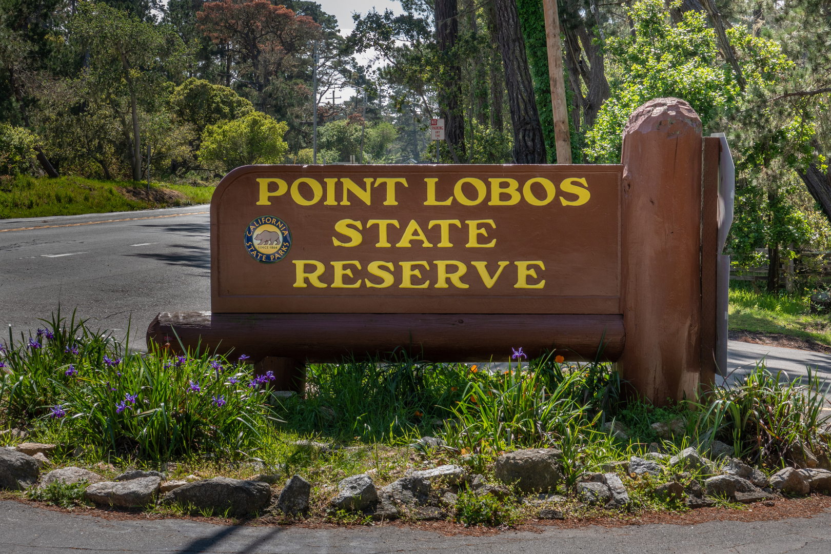 Wooden sign for Point Lobos State Reserve with California State Parks logo, surrounded by flowers and trees.
