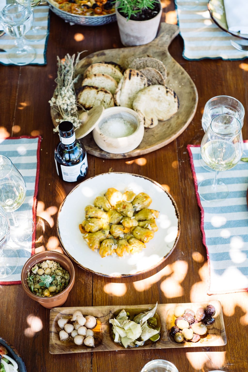 A rustic wooden table set with various dishes including grilled bread, marinated artichokes, a small bowl of hummus, and a plate of stuffed grape leaves, with wine glasses, a bottle of wine or liquor, and small bowls of olives, surrounded by striped 