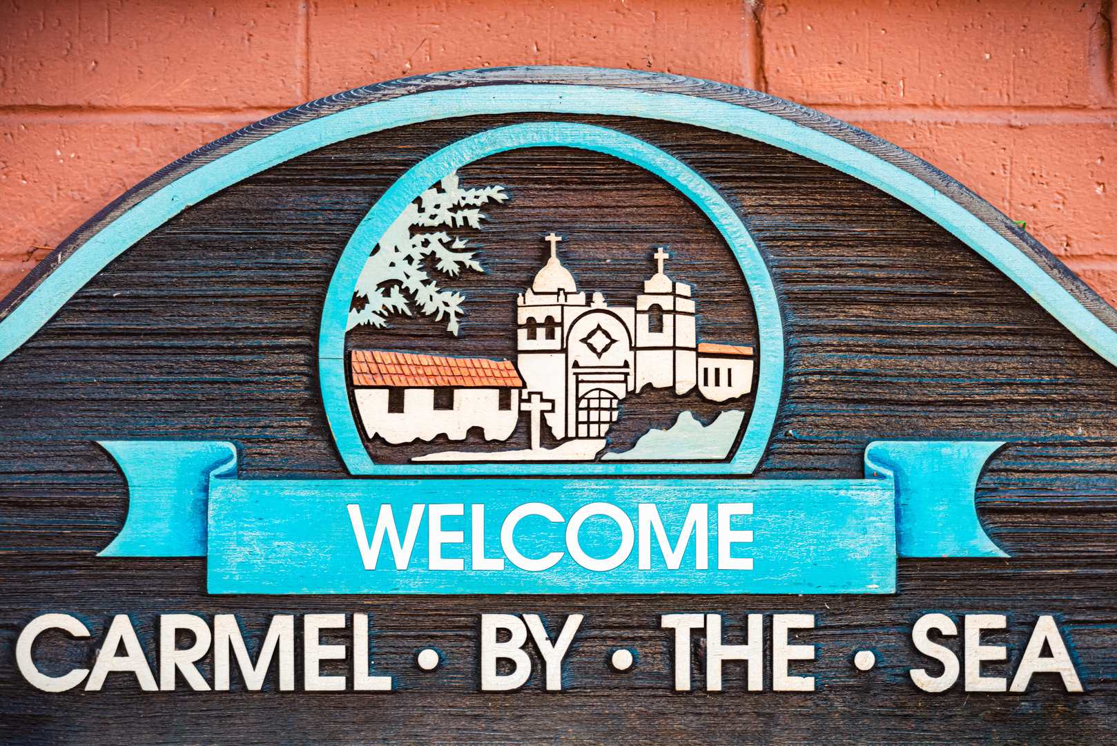 Wooden sign with a cityscape and tree inside a circle, blue banner with 'WELCOME' in white, and partially visible text 'CARMEL BY THE SEA'.