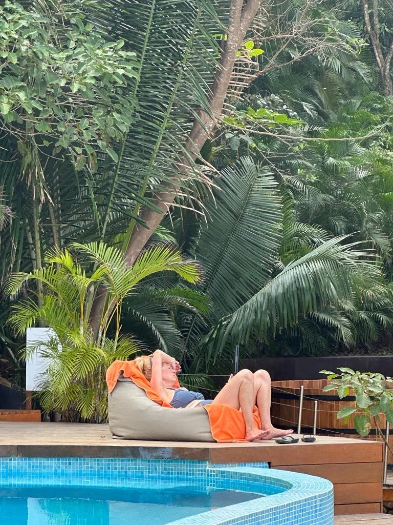 Woman relaxing on a bean bag chair next to a swimming pool, surrounded by lush green tropical plants.