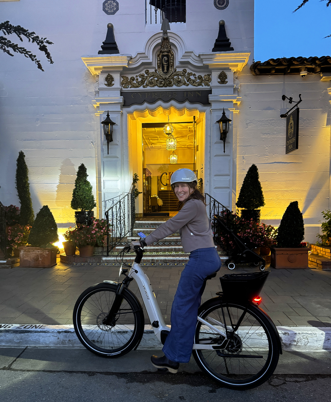 Woman riding an e-bike in front of the Cypress Inn, with the entrance lit up warmly, garden potted plants, and a sign indicating a historic location.