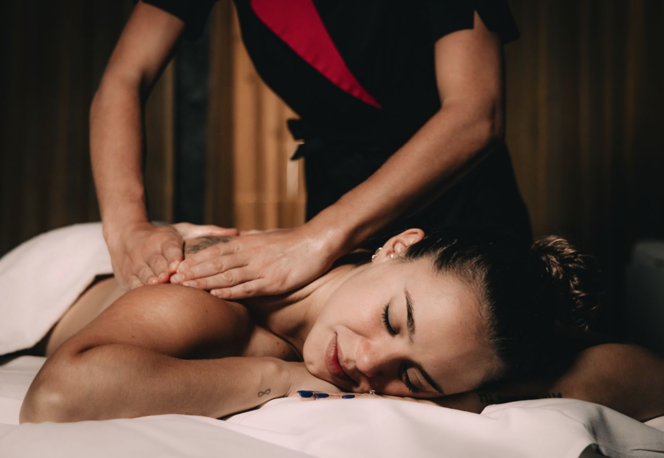 A woman receiving a massage in a spa or massage room, lying face down with eyes closed, while a massage therapist applies pressure to her shoulder area. World Class Spa in Verbier