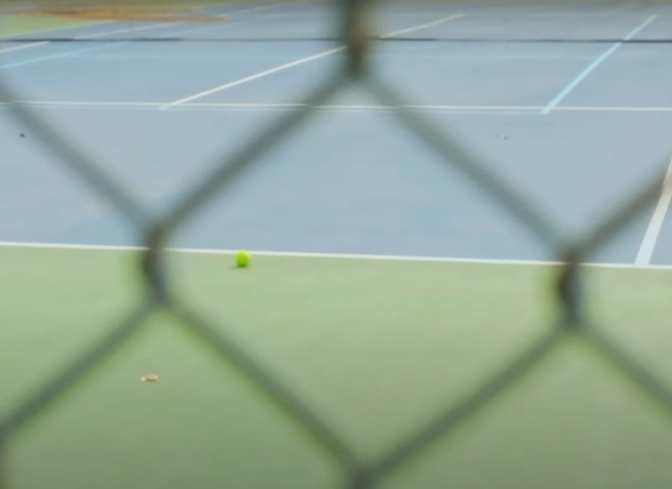 A tennis court seen through a chain-link fence, with a bright yellow tennis ball on the ground in the foreground.