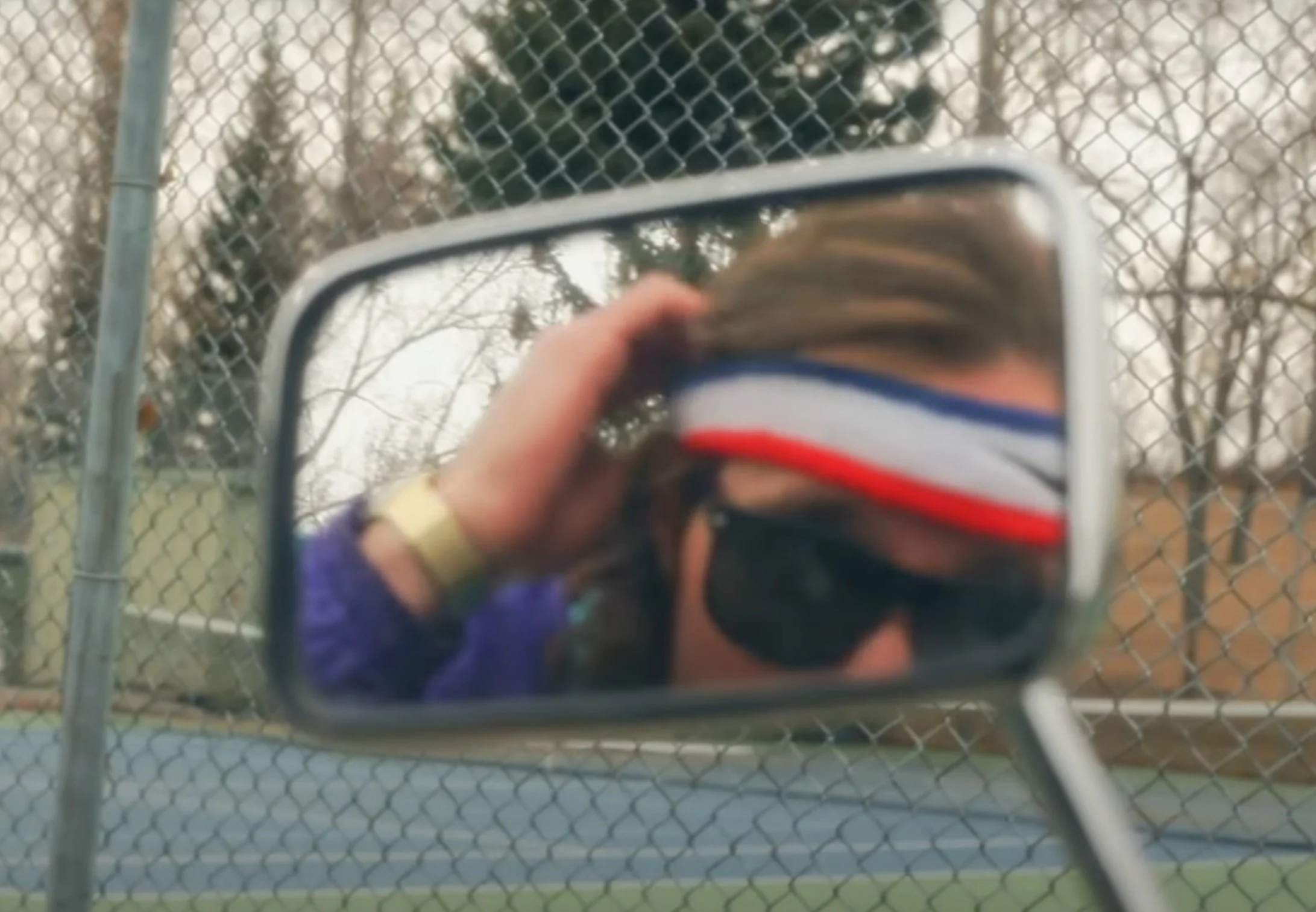 Person taking a selfie in a side mirror wearing sunglasses, a headband with red, white, and blue stripes, and a purple jacket, outdoors near a chain-link fence.