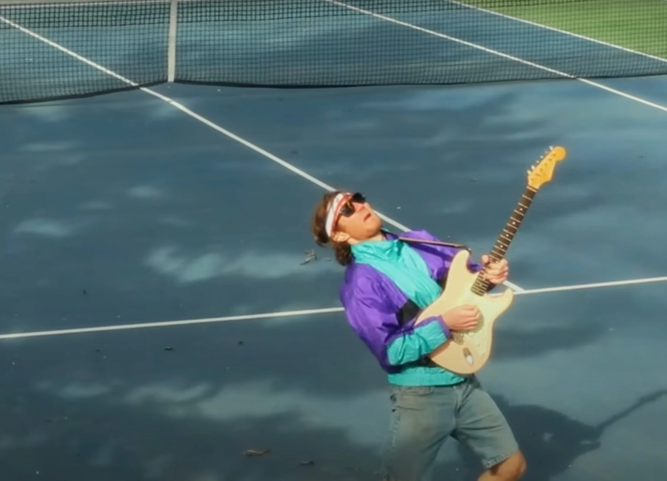 A person wearing sunglasses and a colorful windbreaker jacket is playing an electric guitar on an outdoor tennis court.