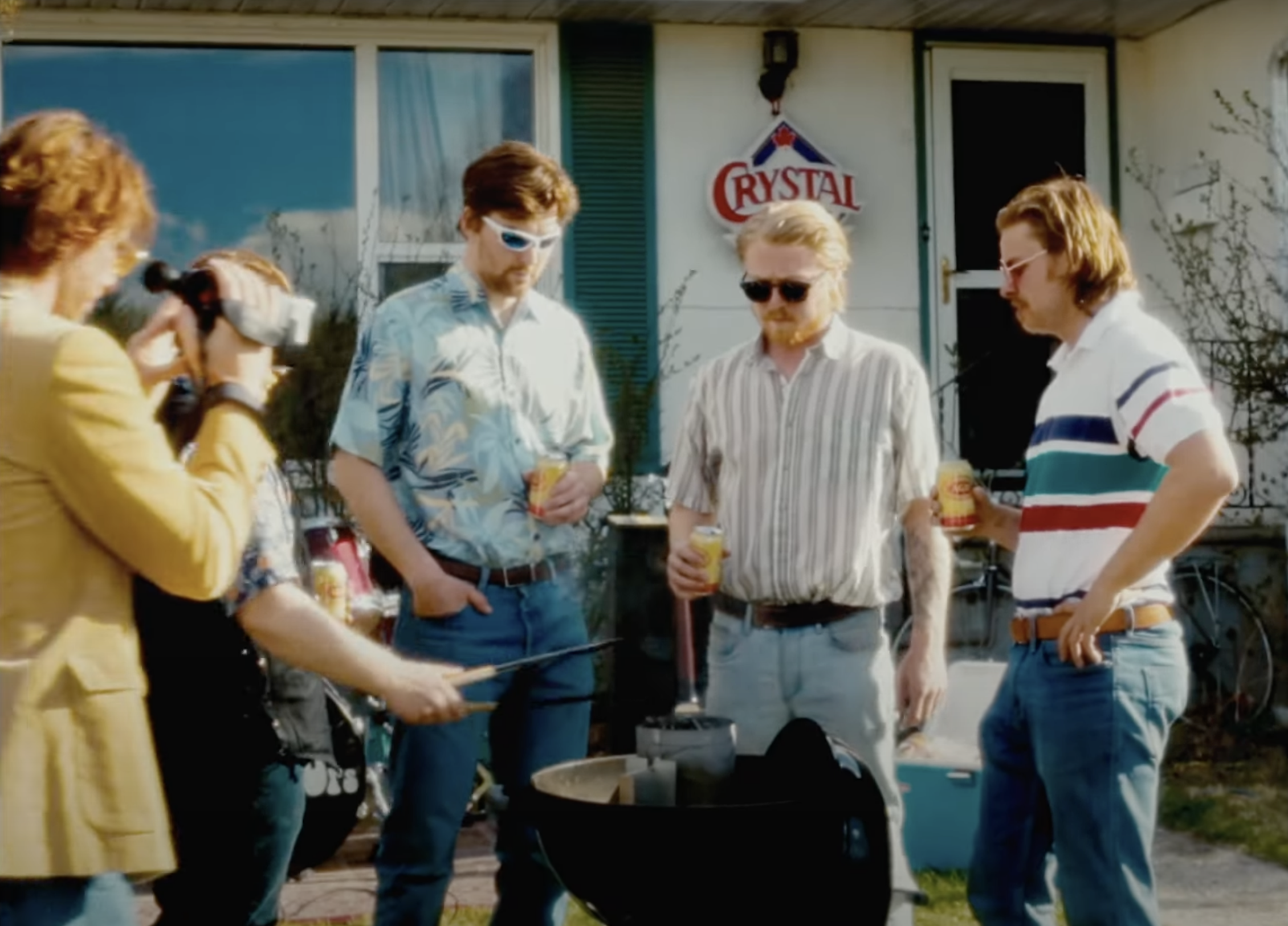 Group of people standing outside, holding drinks, around a barbecue grill, with a woman taking a photo, and a house with a 'Crystal' sign on the wall in the background.