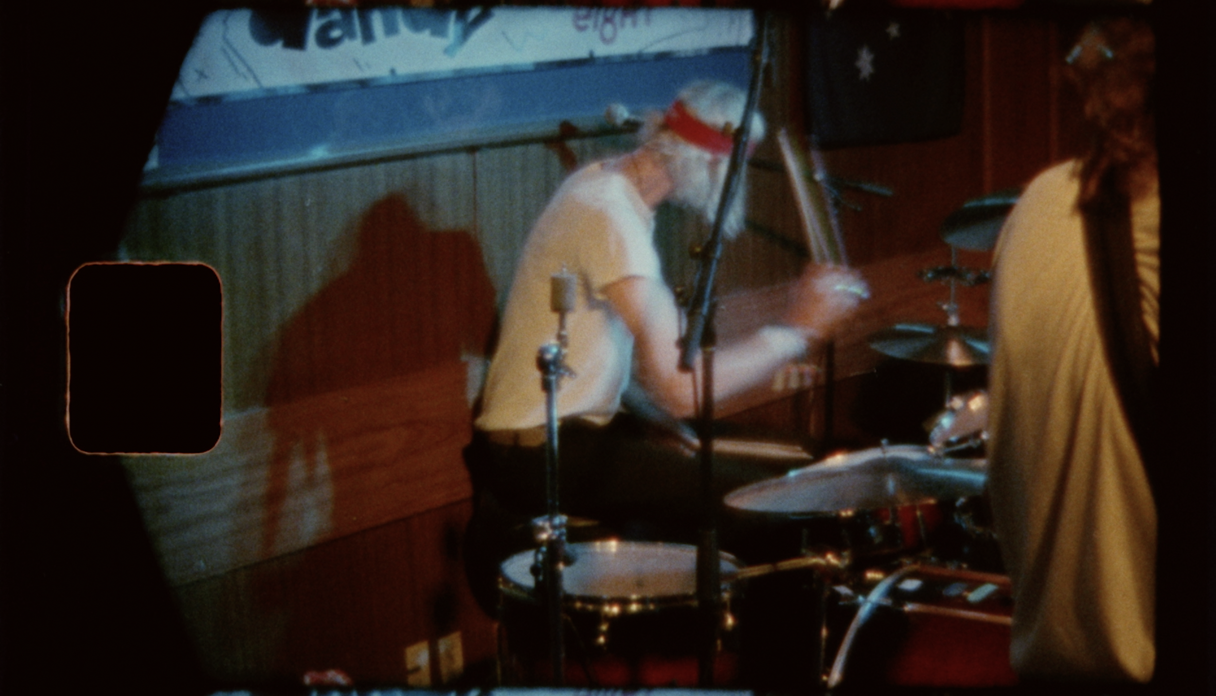 A man with long white hair and a beard, wearing a red headband and white T-shirt, playing drums at a dimly lit indoor venue with wood paneling and a sign in the background.