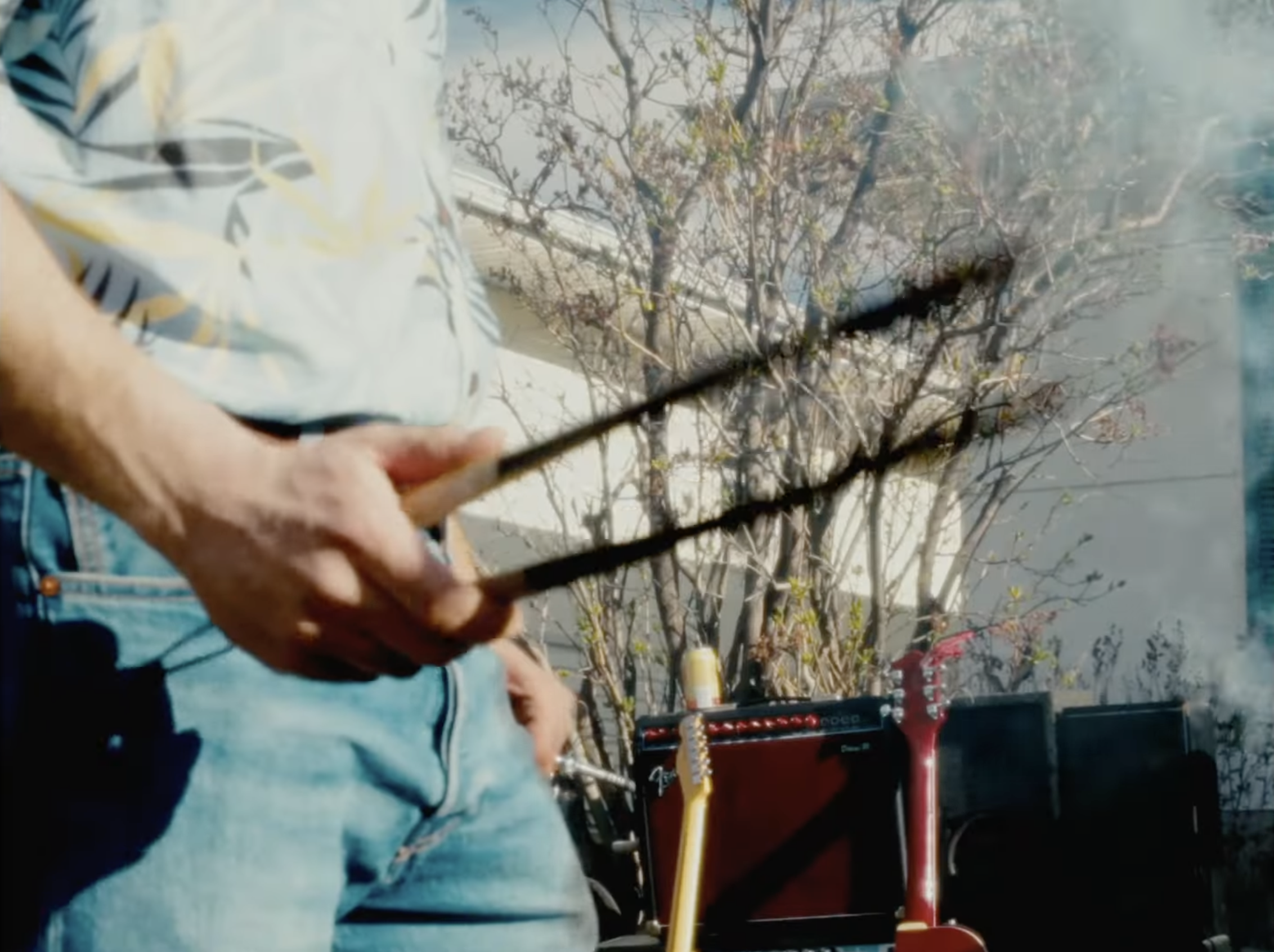 A person wearing a Hawaiian shirt and jeans is holding drumsticks. In the background, there is a small amplifier and two guitars, one yellow and one red, on stands. There are also leafless trees and a building visible behind them.